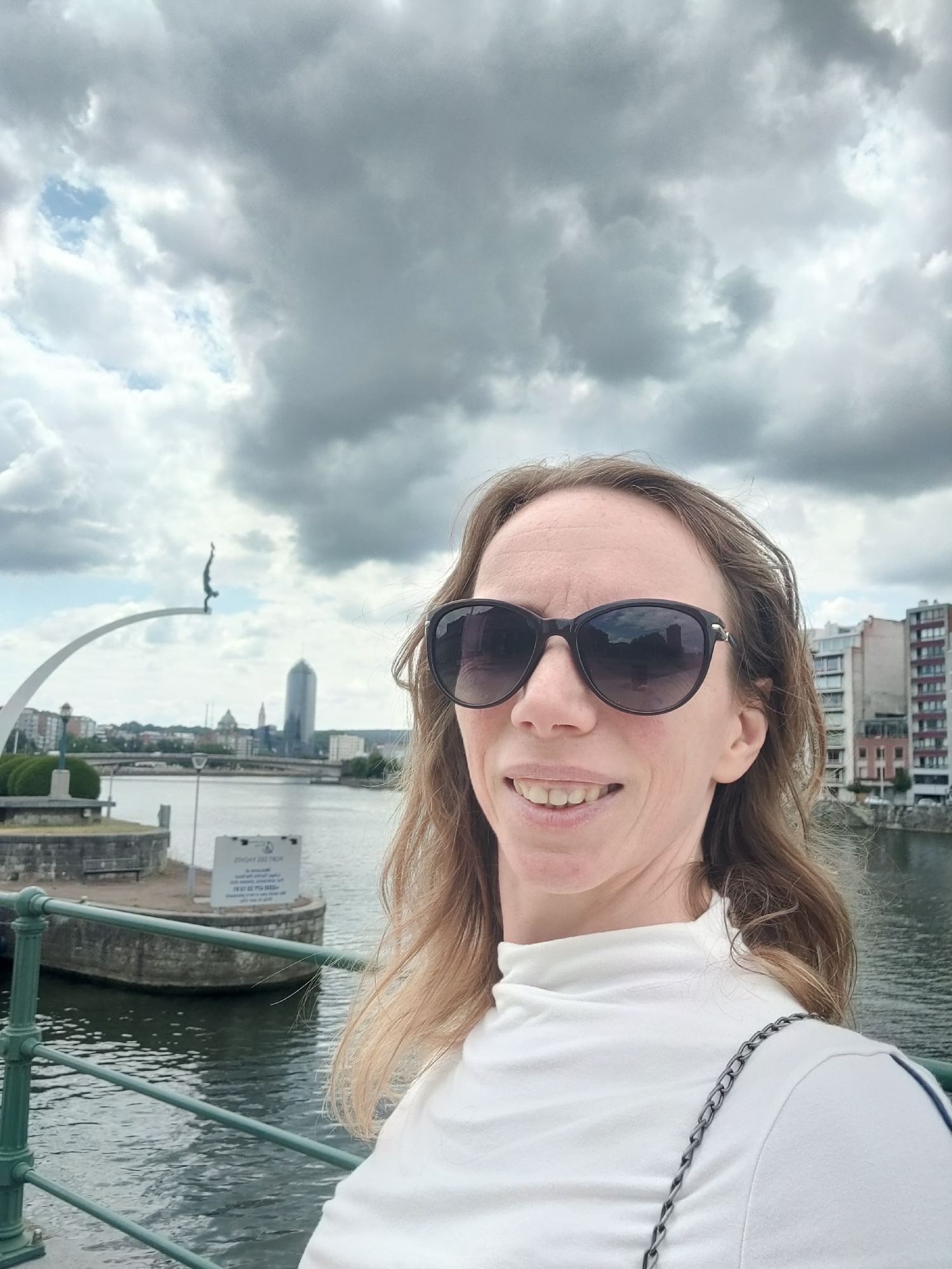 A medium shot captures a woman with shoulder-length brown hair, wearing large dark sunglasses and a white turtleneck shirt. She is standing outdoors, with a body of water and a city skyline in the background. The water appears to be a river, with a green metal railing in the foreground. Behind the railing, a stone embankment is visible, with a bench and a signpost. Further in the background, a modern skyscraper and older buildings line the opposite bank of the river. A sculptural element, resembling a diving figure on an arch, is visible to the left, part of the "Esplanade du Théâtre de la Cascade" in Liège, Belgium. The sky is overcast with dramatic, dark clouds. The woman is smiling, looking at the camera.