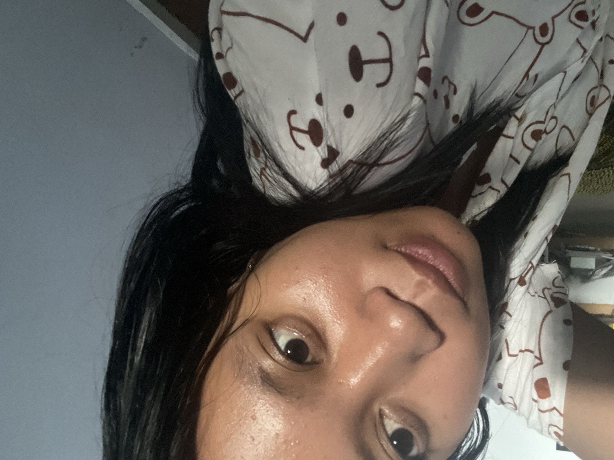 The image is a close-up, sideways selfie of a person in Jambearum, Indonesia. The person has dark hair and is wearing a white garment with a brown bear pattern. The lighting suggests it might be indoors, possibly at night or in a dimly lit room, with a gray wall in the background. The person's face is turned towards the camera, and their expression is neutral. There is no visible text in the image.