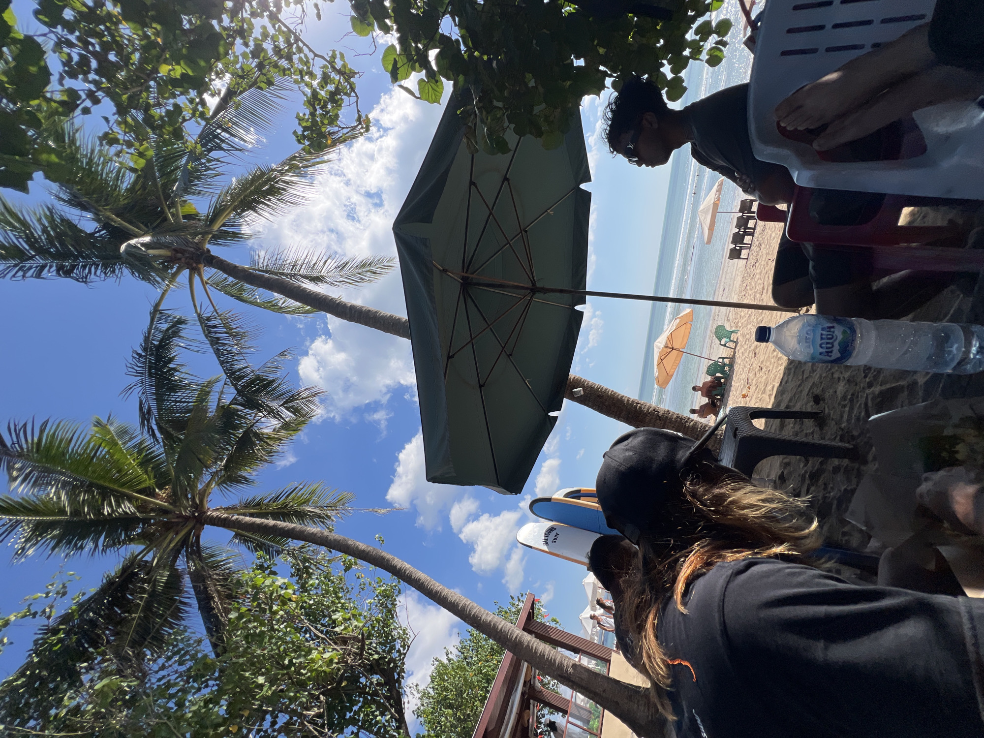 This image depicts a beach scene in Kuta, Indonesia. In the foreground, two individuals are seated on chairs, partially obscured by palm trees and a large dark green umbrella. One person, wearing a black cap and black t-shirt, is facing away from the camera, with their back to the viewer. Their long, blonde-brown hair is visible. The other person, in the middle ground, is facing towards the ocean, wearing sunglasses and a dark t-shirt. Their arms are crossed, and they are seated in a red chair. A partially consumed bottle of Aqua water is placed on the sand in front of them. In the background, several surfboards are propped upright on the beach, along with beach umbrellas and chairs. Other people can be seen in the distance, enjoying the beach and the ocean. The sky is blue with scattered white clouds, and the sun is casting shadows through the palm fronds. The environment is a sandy beach with the ocean and gentle waves visible. The overall impression is one of relaxation and leisure on a sunny day.
