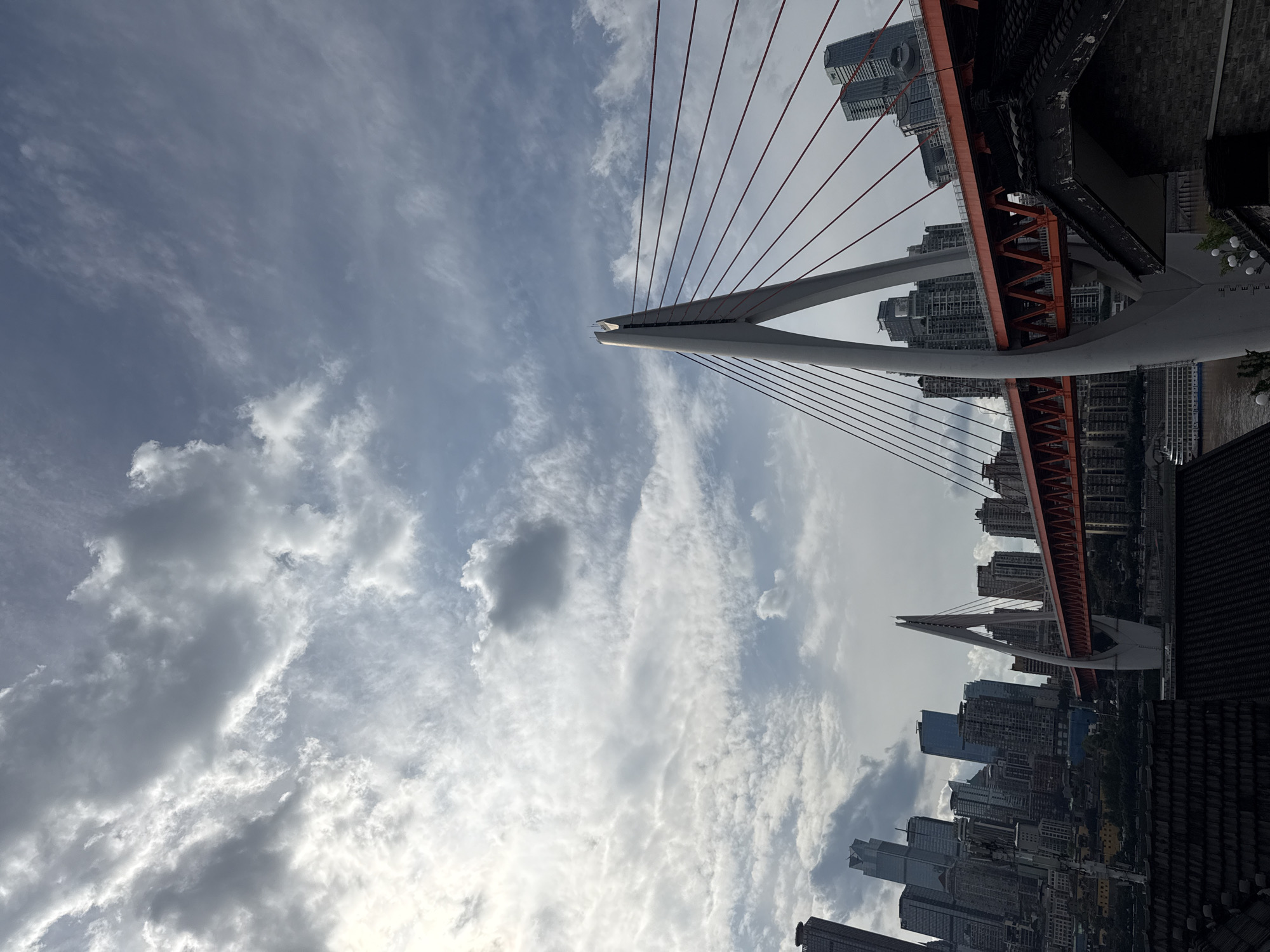 This image captures a dynamic low-angle perspective of a modern cityscape dominated by a large cable-stayed bridge. Two sleek, white A-frame pylons rise prominently into the sky, from which numerous thin, red cables elegantly descend to support the bridge's main structure. The bridge deck itself is a robust orange-red truss, spanning across what appears to be a wide river or waterway visible in the background.
The surrounding area is a dense urban environment, with countless tall buildings and modern skyscrapers forming a bustling city skyline behind and beneath the bridge. The sky is partly cloudy, featuring patches of clear blue, wispy white clouds, and some heavier grey cloud formations, indicating a variable weather day. In the immediate foreground on the right, dark tiled rooftops and decorative street lights suggest a developed pedestrian area or promenade. The scene is brightly lit by daylight, showcasing the grandeur of modern infrastructure and urban development characteristic of a large city like those found in China's Nan'an District. No specific activities or legible text are visible.