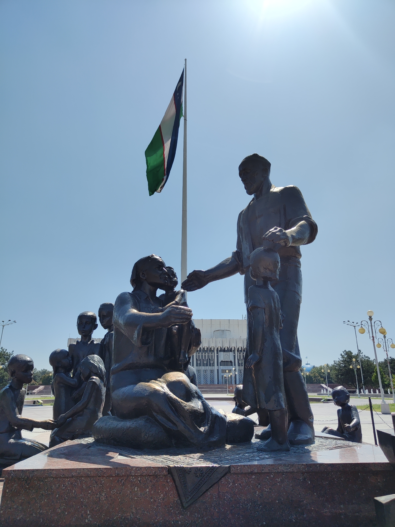 This image depicts the Monument of Independence in Tashkent, Uzbekistan, under a clear blue sky with a bright sun. The monument features a series of bronze statues forming a significant focal point. In the foreground, a large bronze sculpture depicts a male figure standing and gesturing towards a young child, with other figures, including women and children, arranged around them, some seated and some standing. The figures appear to be engaged in communal or familial interactions. Behind the statues, a tall flagpole flies the flag of Uzbekistan, a tricolor of blue, white, and green stripes with a crescent moon and stars. In the background, a large, modern building with a distinct architectural style is visible, along with lampposts and trees, suggesting a public urban space. The base of the monument is a stepped platform with a reddish granite finish. The overall scene conveys a sense of civic pride and national symbolism.