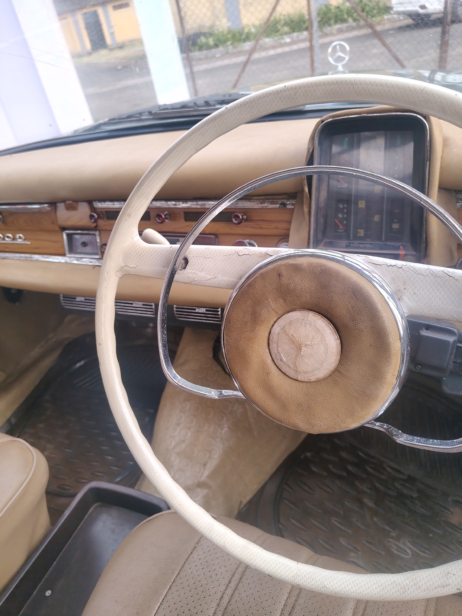 This is a close-up interior shot of a vintage Mercedes-Benz car, likely taken from the driver's seat. The most prominent subject is the cream-colored, two-spoke steering wheel, which shows signs of age with visible cracks and wear on its textured grip. The center of the steering wheel features a tan leather-wrapped horn pad, encircled by a chrome ring, with a lighter, circular insert at its core that appears to have a star-like pattern.
The dashboard behind the steering wheel is predominantly tan, with a wooden trim panel across its lower section. This wooden panel houses various buttons, switches, and air vents. Partially visible on the left side of the dashboard's wooden trim is the text "Pood". Behind the steering wheel, a rectangular instrument cluster shows a speedometer with readings up to 80, marked with numbers 20, 40, and 60.
Through the windshield, a clear Mercedes-Benz star emblem is visible on the hood of the car, identifying the vehicle's brand. The outdoor background is blurred but shows what appears to be a building with a yellow-brown wall, a chain-link fence, and some greenery, indicating a daytime setting.
The car's interior also features light tan/beige upholstered seats, with perforations visible on the passenger seat. The floor is covered with dark, textured mats. There are no people visible in the image. The scene suggests the car is stationary. The provided location context is Kalamassery, India.