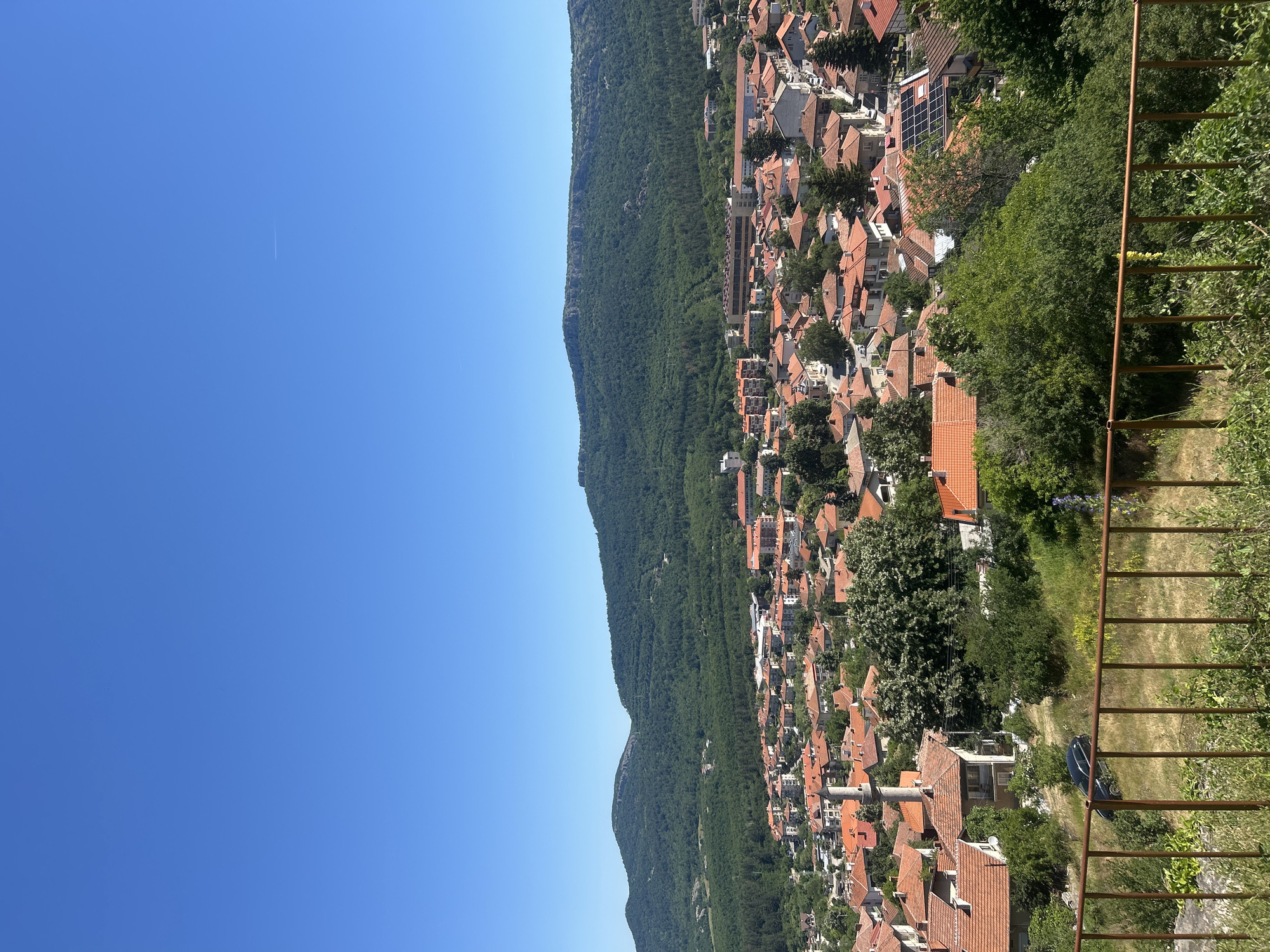 The image shows a panoramic view of the city of Belogradchik, Bulgaria, nestled against a backdrop of lush green hills. The foreground features a rusty metal fence, with dry grass and greenery below. Beyond the fence, the town unfolds, characterized by numerous buildings with distinctive red-tiled roofs. A tall, slender minaret, typical of a mosque, stands prominently among the rooftops. In the distance, verdant hills rise, their slopes densely covered with trees. The sky is a clear, vibrant blue, with a faint contrail from an airplane visible in the upper portion. The overall scene depicts a sunny day in a European town.