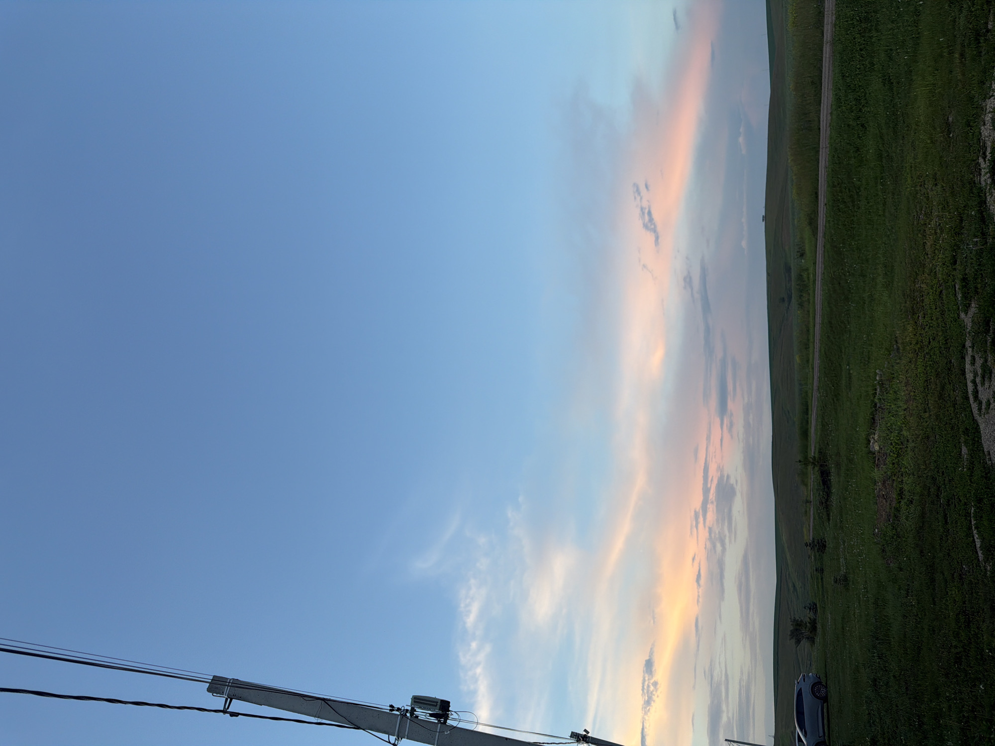 The image depicts a rural landscape under a twilight sky, not in the city of Nizhny Suyk-Su, Russia. A utility pole with wires is prominent in the left foreground, angled towards the sky. Beyond the pole, a light-colored SUV is parked on a grassy area. The midground features rolling green hills, likely farmland, with a dirt road traversing them. The sky transitions from a deep blue at the top to softer hues of pink and orange near the horizon, indicating the sun has recently set. Wispy clouds are scattered across the sky, catching the fading light. The overall scene is one of quiet, natural beauty in a rural setting.