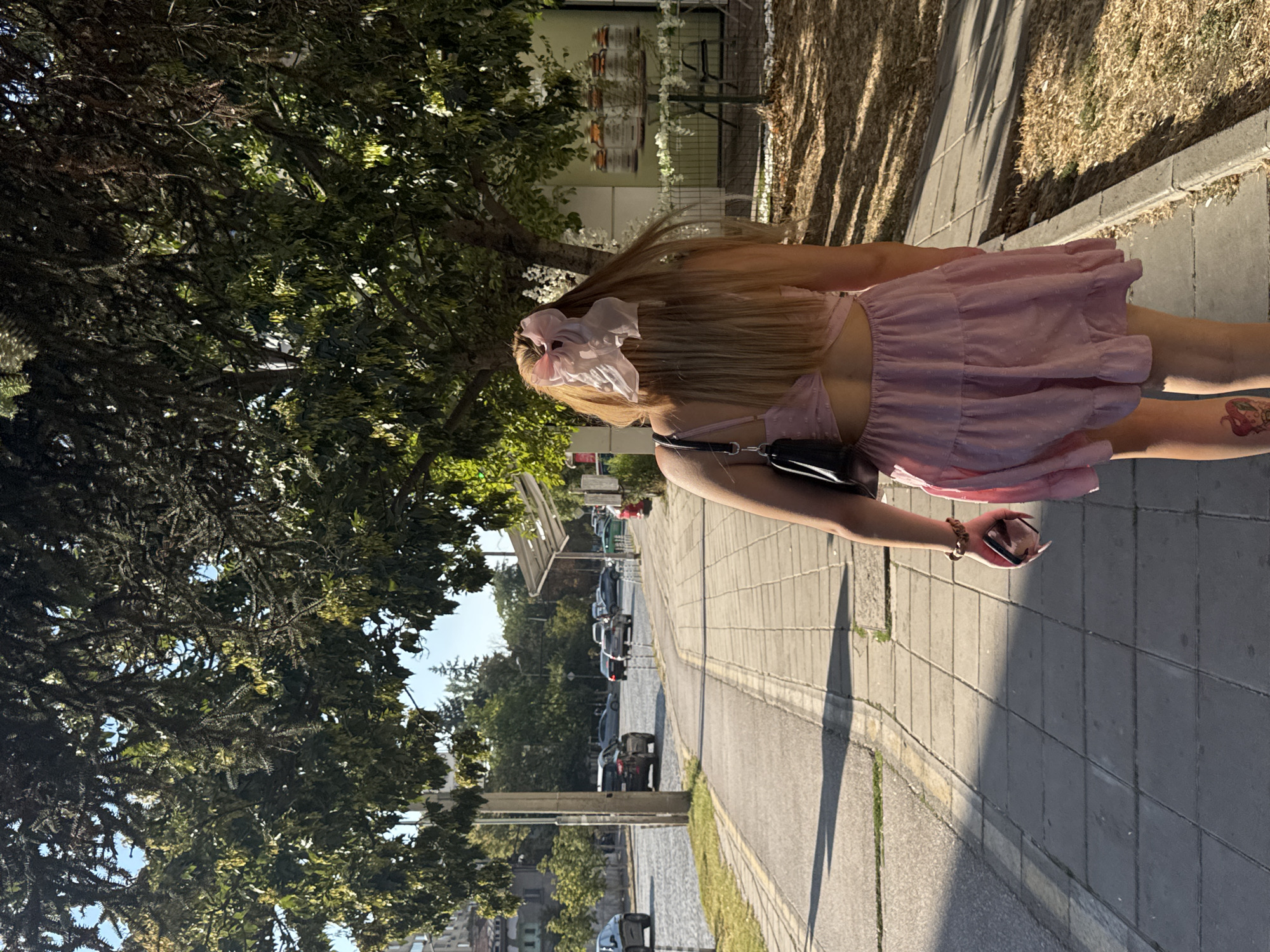 A young woman with long blonde hair, adorned with a large pink scrunchie, walks away from the camera down a paved sidewalk in Sofia, Bulgaria. She is wearing a pink, ruffled, sleeveless dress and carrying a black purse on her left shoulder. In her right hand, she holds a mobile phone. Sunlight casts long shadows from trees overhead onto the sidewalk. The street, with cars in motion, runs alongside the sidewalk on the left. Beyond the street are buildings and more trees. To the right of the sidewalk is a grassy area.