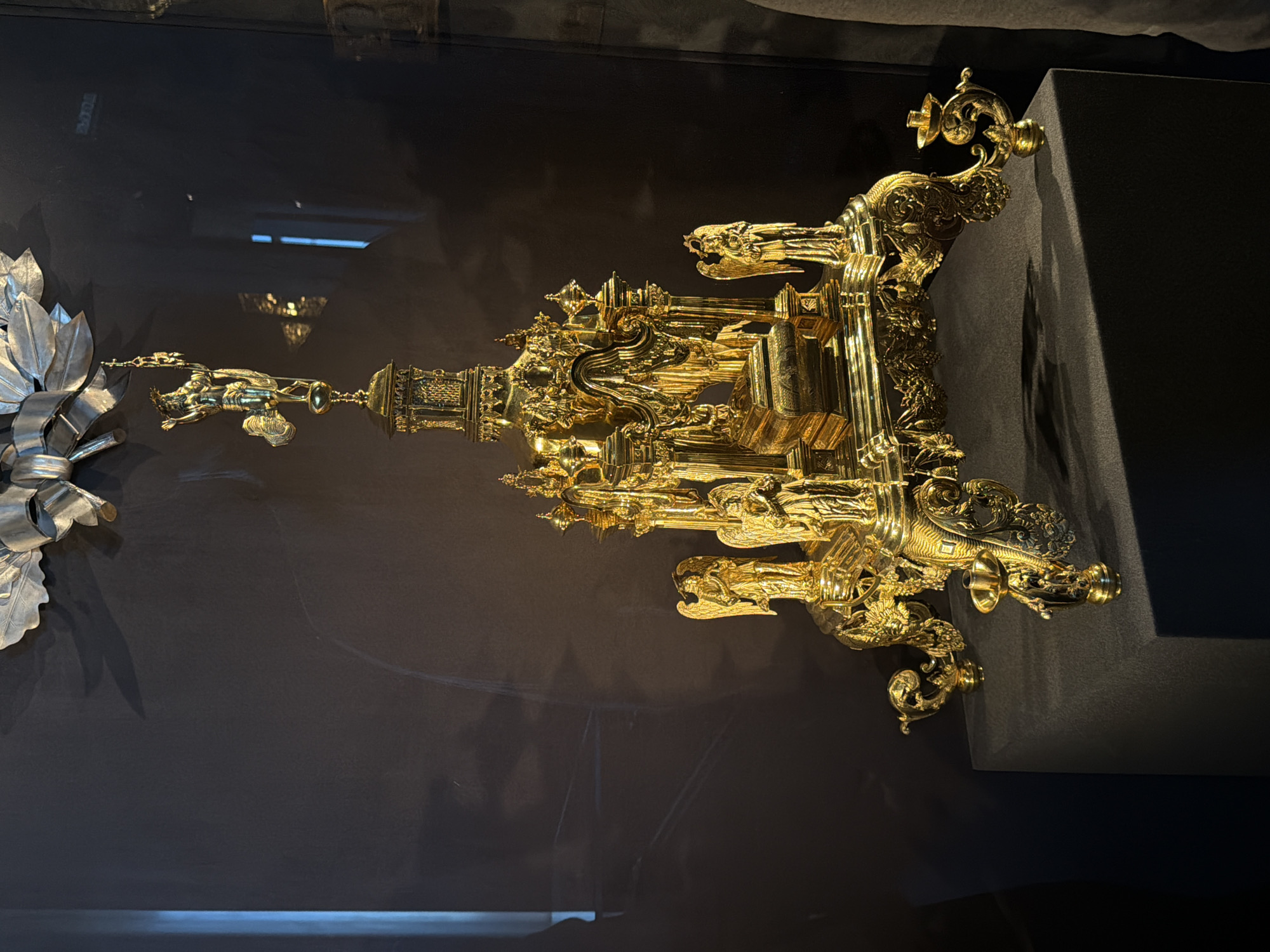 A close-up, slightly angled shot depicts a gold-colored, intricately designed artwork or sculpture displayed in what appears to be a museum or gallery setting. The sculpture rests atop a dark, possibly rectangular stand. The background consists of a smooth, dark-colored wall. The overall environment suggests a controlled, indoor setting designed for showcasing art pieces. There is no mention of Perm or Russia and no people present in the shot.