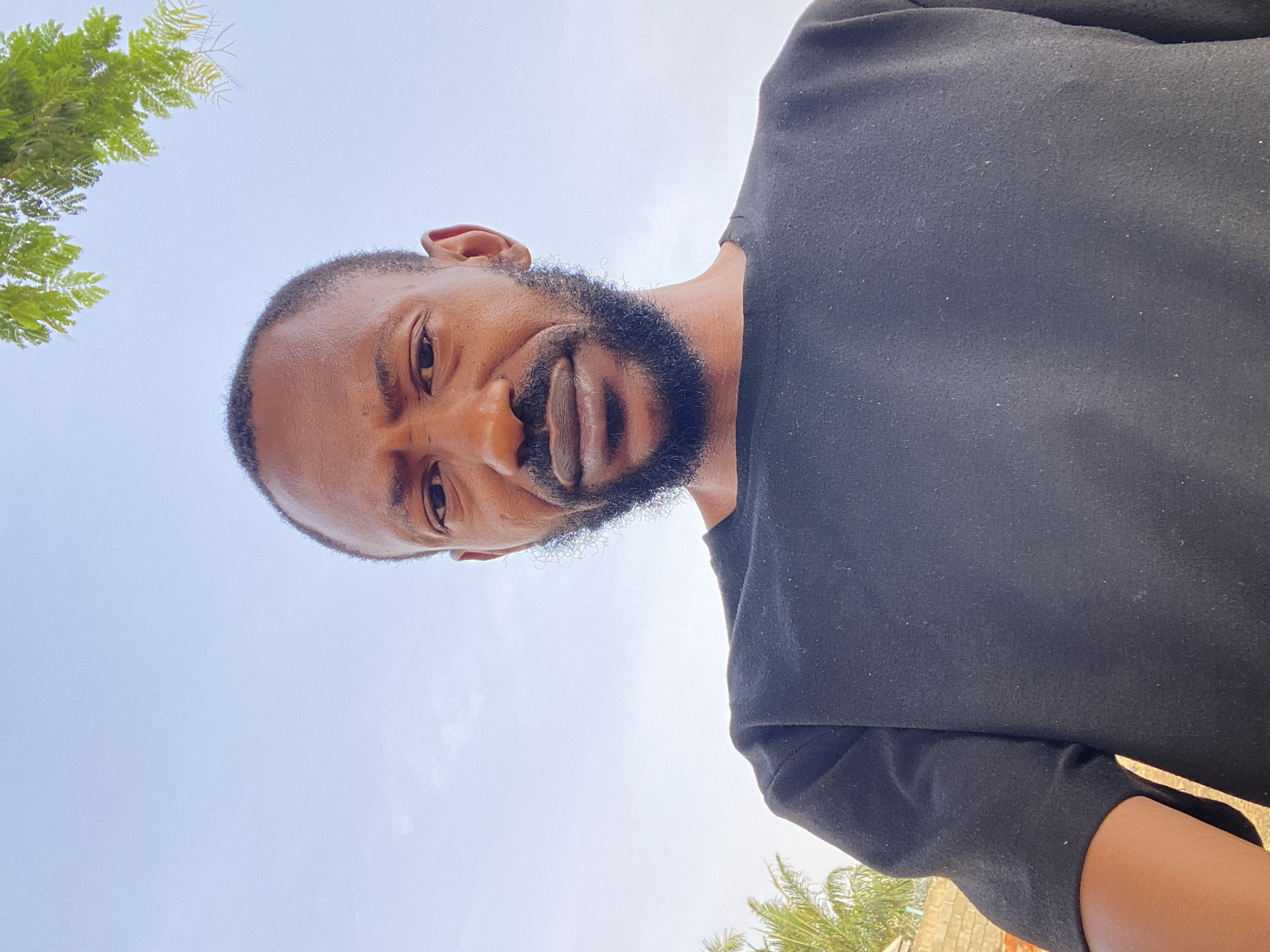 A man with a beard and short hair looks down at the camera. He wears a black t-shirt. The background consists of the sky, some trees, and a building. The location is Sabon Tasha, Nigeria.