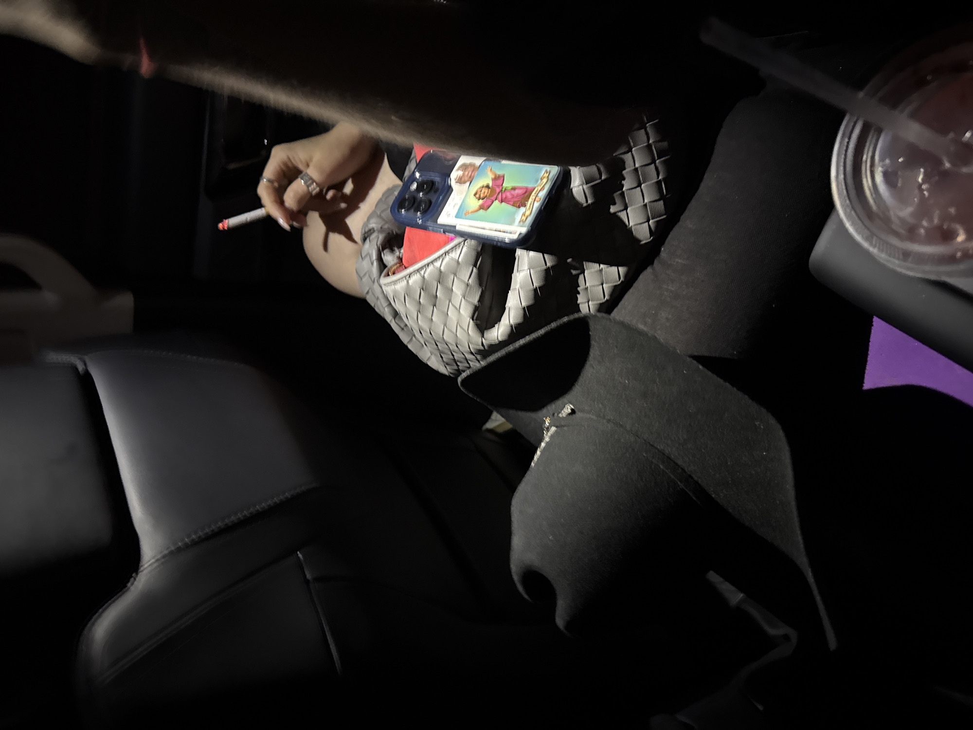 The image shows the interior of a car. A hand holds a lit cigarette. On the car seat rests a grey woven bag with a cell phone on top. The cell phone has a religious image affixed to it. Next to the bag sits a cup with a straw. A dark hat is next to the cup. The lighting suggests nighttime. There is no information in the image indicating that this location is in the city of Encarnación de Díaz, Mexico.