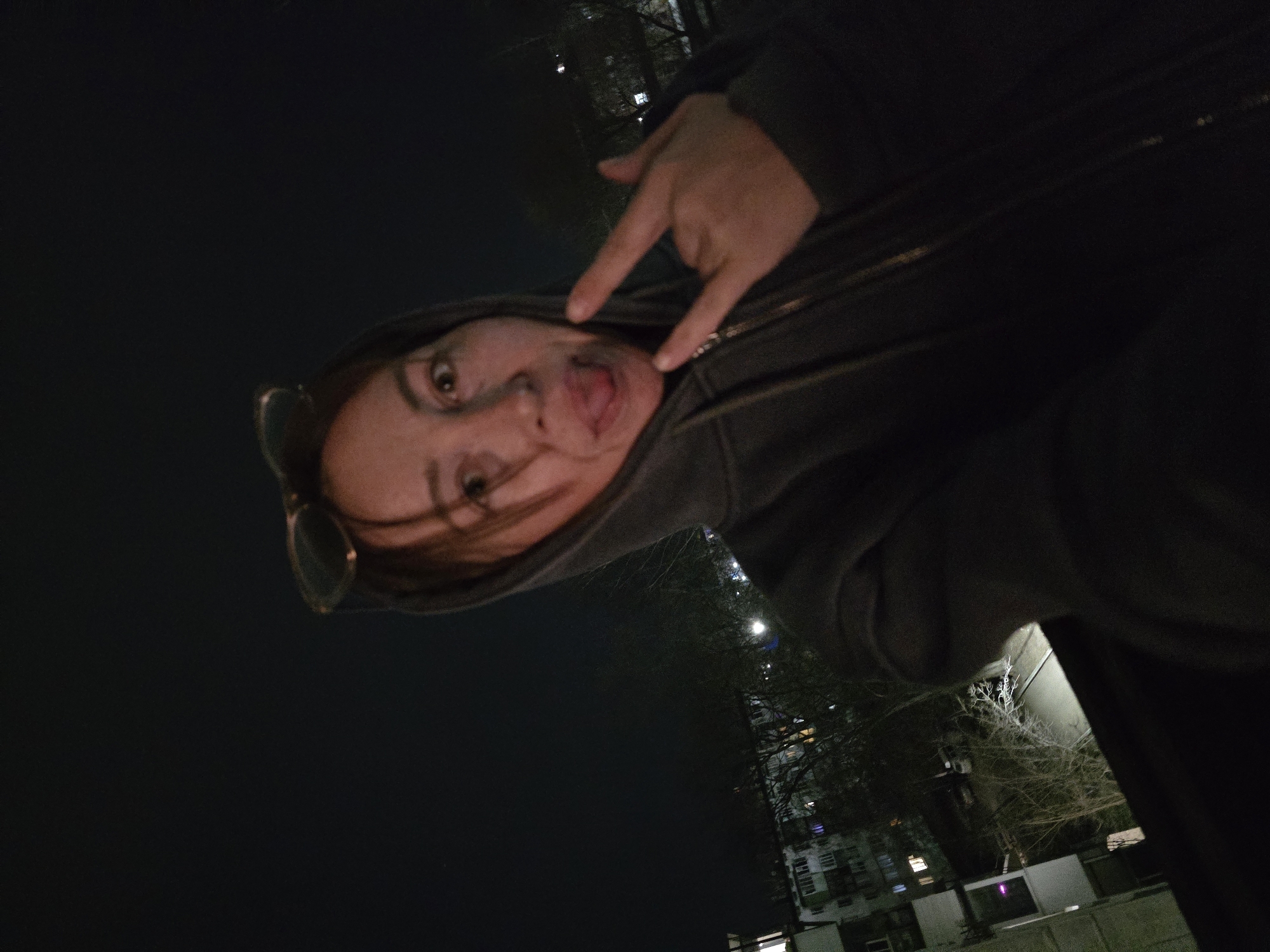 A person, presumably female based on facial features, is making a "rock on" hand gesture while sticking out their tongue. The person is wearing a hooded jacket with sunglasses perched on their head. The photograph appears to be taken outdoors at night with building lights visible in the background, suggesting an urban environment. The image contains trees, a multi-story building and street lights in the background. The approximate location is the city of Chaikovsky, Russia, but the specific street address or landmark is not discernible from the image.