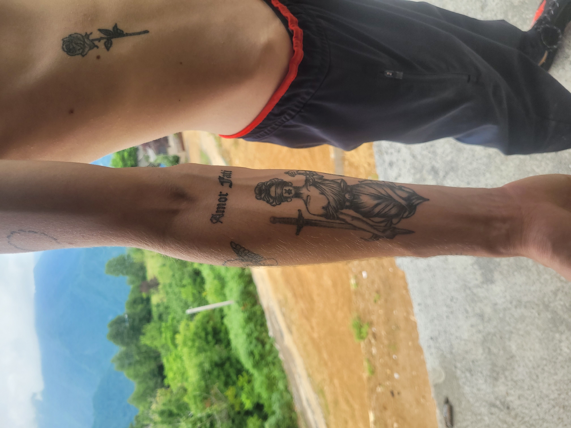 A man's forearm, tattooed with the words "Amor Fati" and an image of Lady Justice holding a sword and scales, is positioned in the foreground. A rose tattoo is visible on his rib cage. He is wearing dark blue pants with a red waistband. The background features a mountainous landscape with lush green trees and a cloudy sky. The scene is outdoors, with a concrete surface in the lower portion of the frame. The location is identified as Milisi, Georgia.