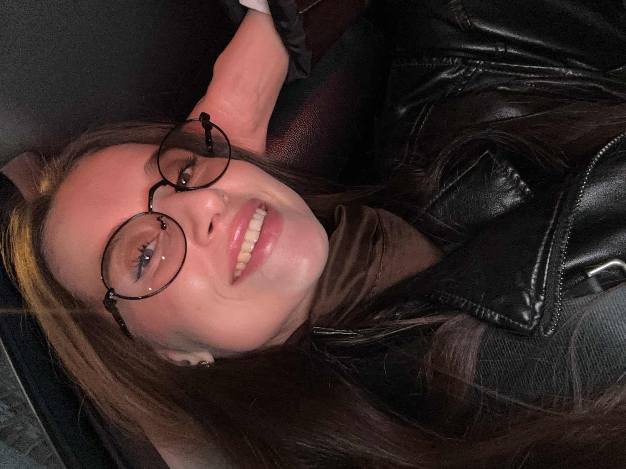 A woman wearing round glasses is lying on her side, smiling. She is wearing a brown turtleneck sweater and a black leather jacket. The background is dark. The location cannot be determined from the image.