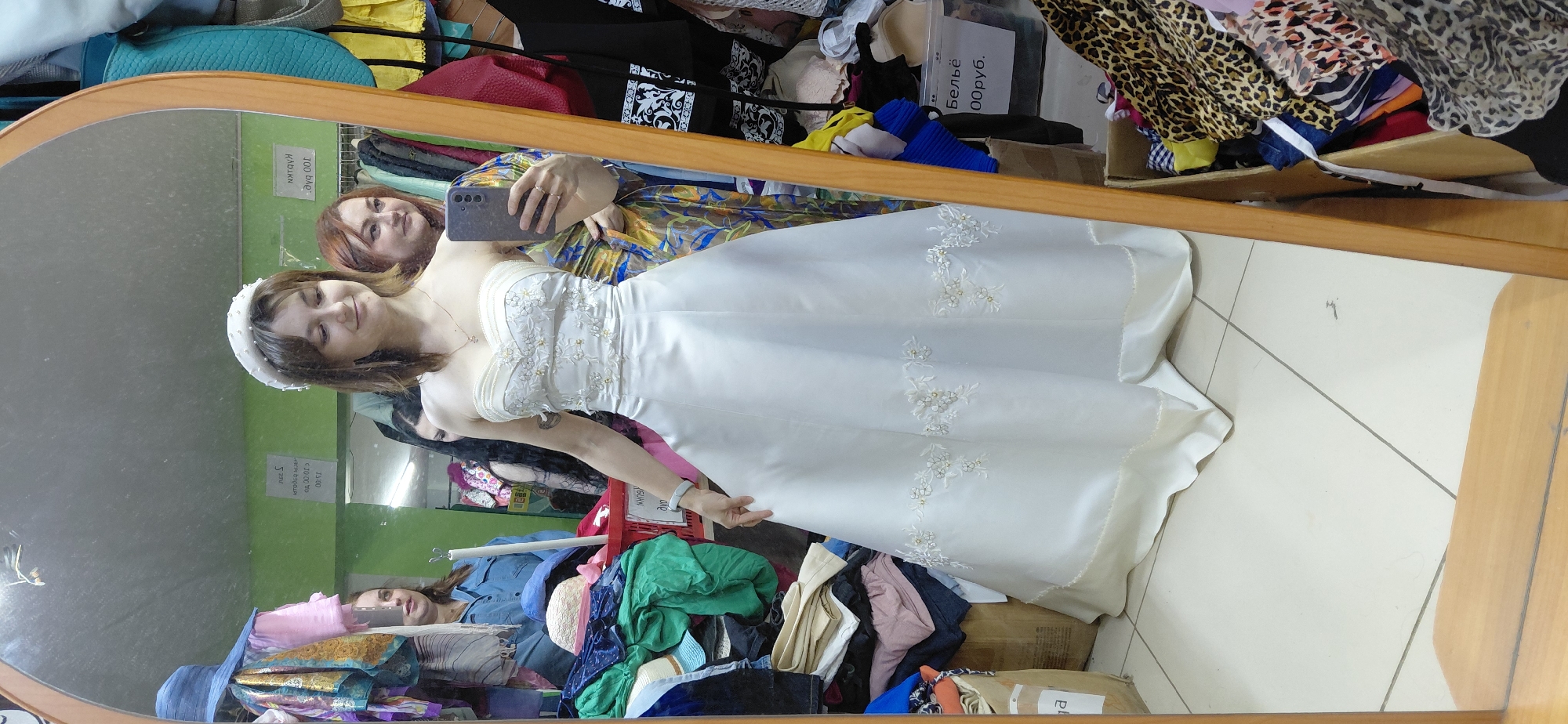 In Magnitogorsk, Russia, a young woman is captured in a full-length mirror reflection, trying on a strapless white wedding dress. The dress features a fitted bodice adorned with intricate white floral embroidery and delicate beading, transitioning into a full A-line skirt. She wears a simple white beaded headband. To her right, another woman with auburn hair looks on, holding a smartphone, possibly taking a photograph. In the background, to the left of the bride, another woman in a blue denim shirt and a blue wide-brimmed hat is visible. The environment appears to be a retail space, possibly a second-hand clothing store, with racks of clothing and stacks of clothes in boxes filling the background. A price tag reading "Бельё 100руб." (Lingerie 100 rubles) is visible on a box in the lower right corner. The floor is tiled, and the mirror has a light brown wooden frame.
