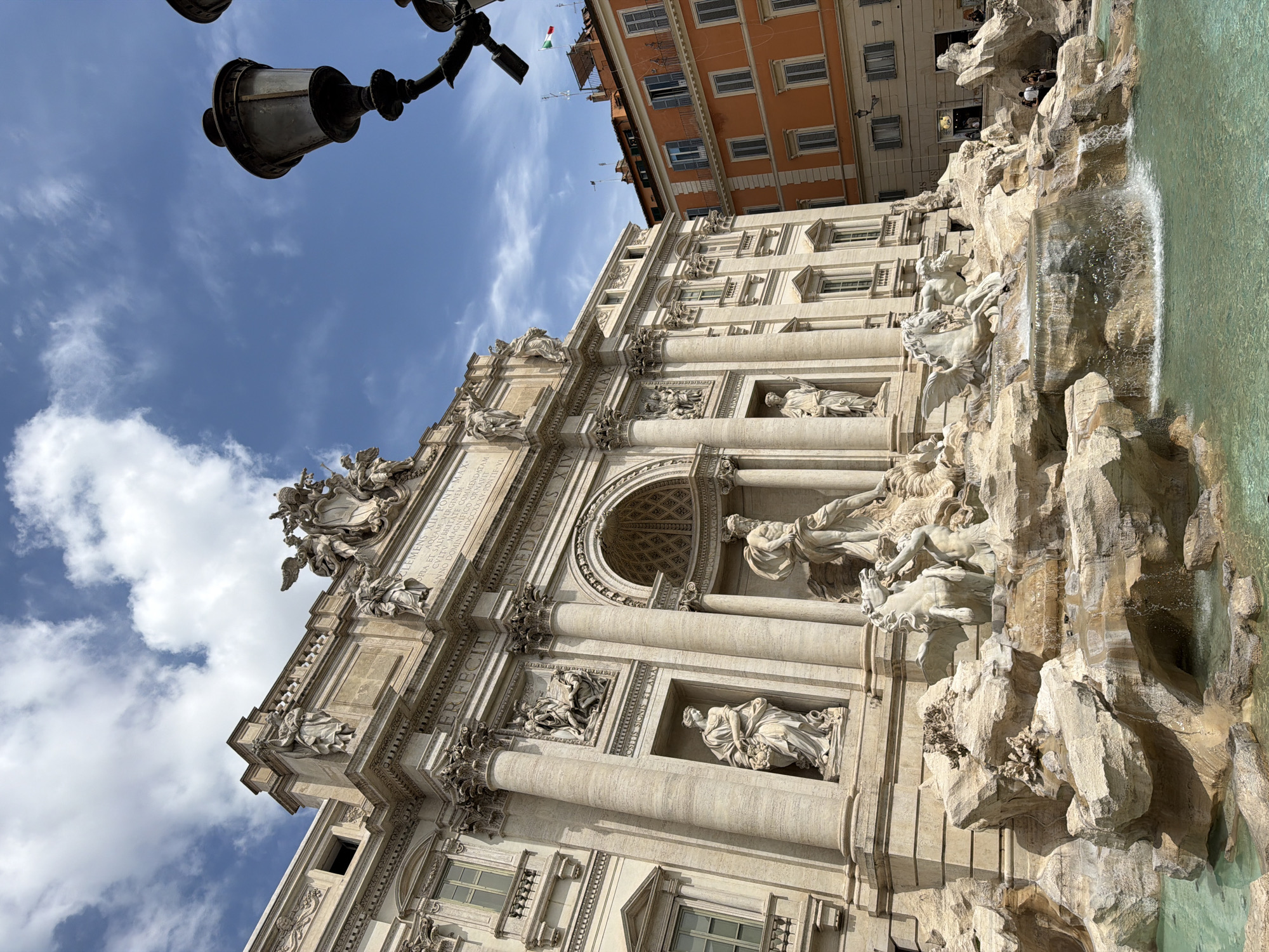 This low-angle photograph captures the majestic Trevi Fountain in Rome, Italy, under a bright, partly cloudy sky. The grand Baroque architecture of the fountain dominates the scene, featuring numerous detailed sculptures and classical elements. At the center, a large statue of Oceanus (Neptune) stands beneath a triumphal arch, flanked by allegorical figures and Tritons guiding seahorses. Water dramatically cascades over the rugged, rock-like formations into the turquoise-green basin of the fountain.
The perspective looks upwards, revealing a clear blue sky with scattered white clouds. An ornate, dark lamppost extends into the upper left corner of the frame. In the background, above the fountain's facade, parts of adjacent buildings are visible, including one with an orange facade on the right, from which a small Italian flag (green, white, and red) flies. Several small, indistinct figures of people can be seen on and around the rocky structure of the fountain and near the buildings in the background, suggesting a typical day at a popular landmark.
Notable Latin inscriptions are visible on the fountain's facade, including "CLEMENS XII PONT. MAX. AQVAM VIRGINEM CVLTV MAGNIFICO ORNAVIT ANNO DOMINI MDCCXXXV PONTIF VI PERFECIT". These texts commemorate Pope Clement XII and the fountain's construction. The bright lighting indicates it is daytime, likely late morning or early afternoon.