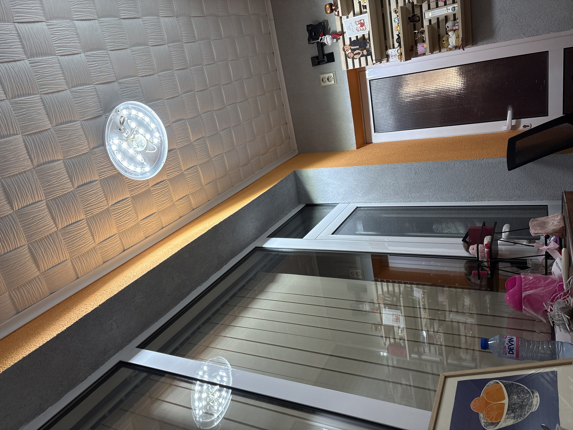 The image depicts the interior of a room with a textured, possibly woven, pattern on the ceiling and an LED light fixture attached. Below the ceiling is a wall, the upper part of which is painted in an orange color, with a gray color on the lower part. There's a decorative display cabinet or shelf on the wall, holding small figurines and decorations. A window is visible, reflecting an exterior view, and there are some items on the windowsill, including a water bottle and a framed picture featuring oranges. The location in Panagyurishte, Bulgaria, cannot be directly determined from the given image.