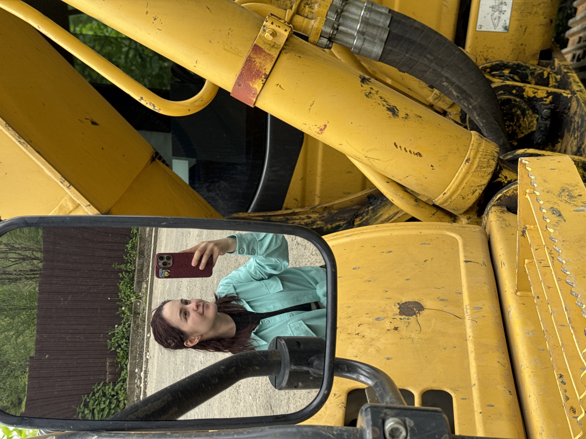 A person is taking a selfie, as reflected in the mirror of a yellow construction vehicle. The location outside the mirror appears to be a street or yard. The city or country is undetermined based on the image.