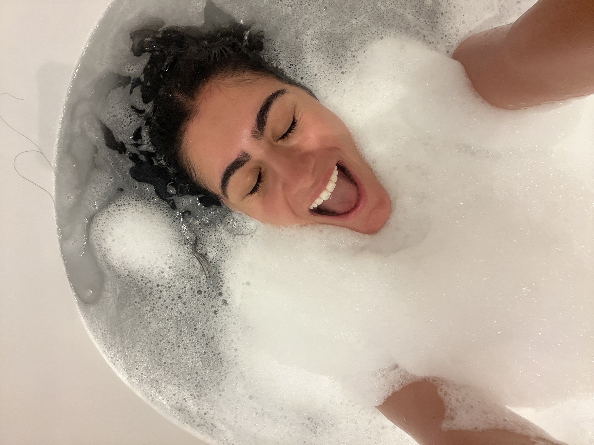 A woman is taking a bubble bath. She is submerged in the water, with only her head and upper body visible. She has dark, curly hair and her eyes are closed. Her mouth is open in a wide smile, and her teeth are visible. Bubbles surround her face and chest. A few strands of hair are visible floating on the surface of the water, and a long strand of dark hair is trailing over the edge of the bathtub, against a light pink background. The bathtub is white and appears to be in a bathroom. There are no explicit indications of the location being Almancil, Portugal, as the image only shows a bathtub and its occupant.