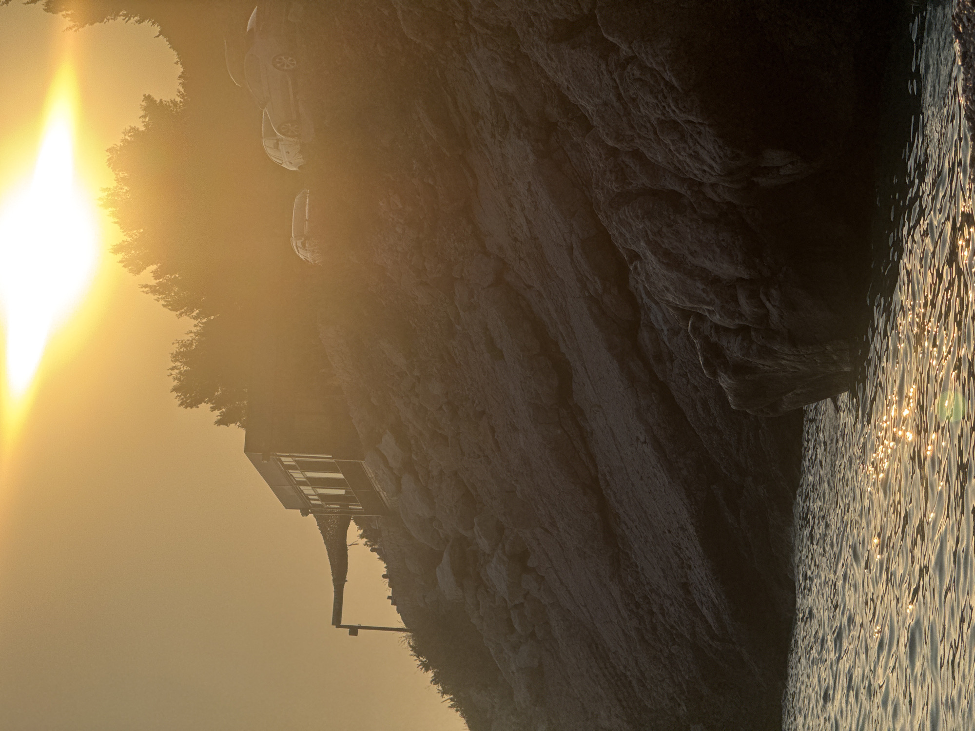 The image depicts a sunset scene in Kamenovo, Montenegro, featuring a rocky coastline and the Adriatic Sea. In the midground, a building with large windows, possibly a restaurant or cafe, is situated on a rocky incline. Several cars are parked on the slope above the building, partially obscured by trees and the strong backlight from the setting sun. The sun itself is a bright, hazy orb in the upper sky, casting a golden glow over the entire landscape and creating shimmering reflections on the water's surface. The foreground shows the gentle waves of the sea lapping against a pebbly shore. The overall atmosphere is serene and bathed in the warm light of dusk.