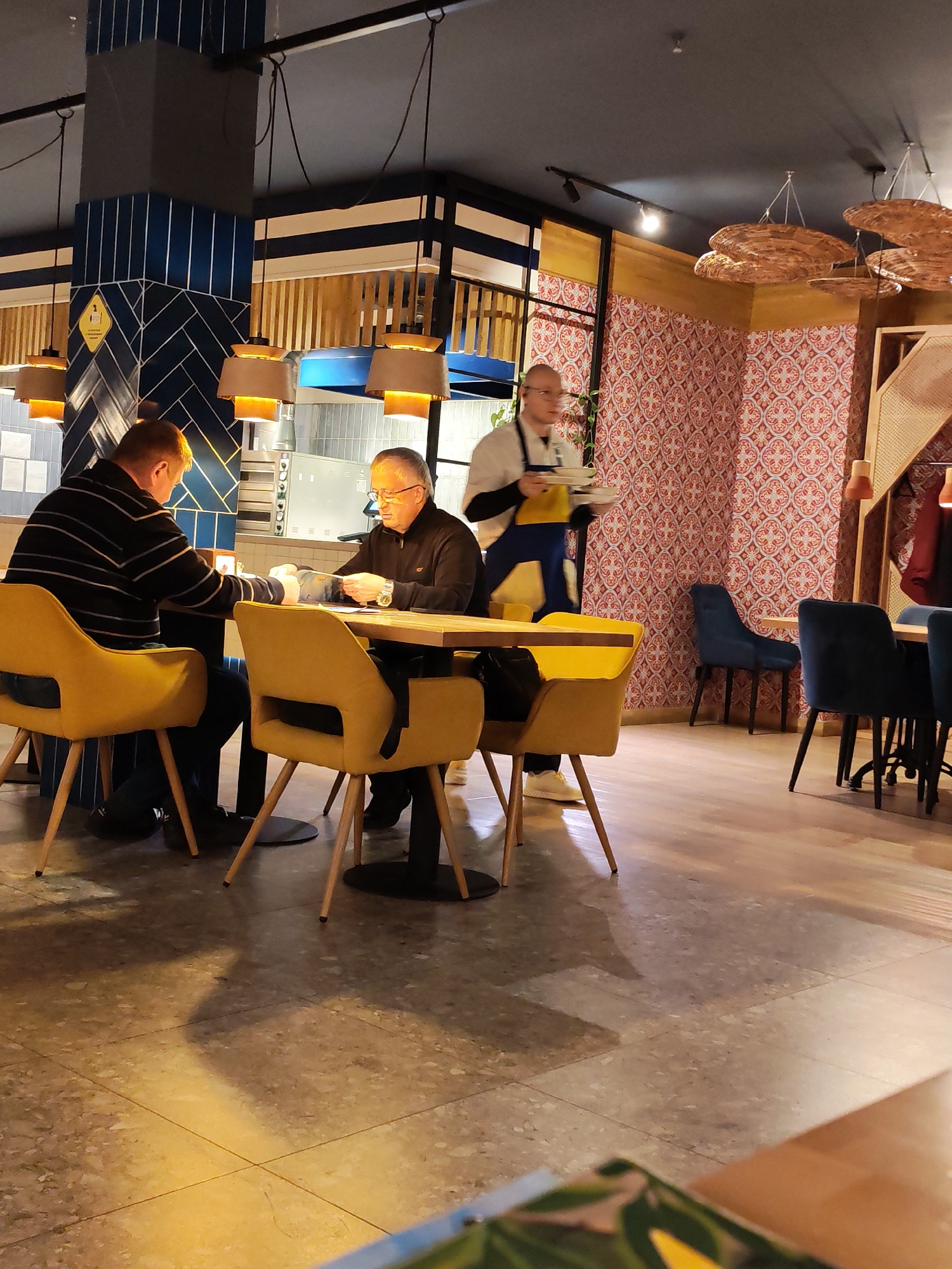 The image captures the interior of a contemporary restaurant or cafe, likely during the evening due to the reliance on artificial lighting. The setting features a mix of textures and colors, including a prominent column tiled with dark blue patterns – herringbone at the bottom and vertical stripes above. A yellow sign with a "2" symbol is visible on this column, possibly indicating a social distancing measure.
In the mid-ground, two men are seated at a wooden table with modern, mustard-yellow upholstered chairs. The man on the left, seen from behind, wears a striped long-sleeve shirt. The man opposite him, an older individual with glasses and a dark top, appears to be examining something in his hands, possibly a menu or documents.
To the right, a staff member, also wearing glasses and dressed in a white chef's-style top with a blue apron, is carrying a stack of white plates. He is in motion, walking past the seated customers.
The background on the right features a distinctive wall covered in patterned wallpaper, characterized by red and white/light blue geometric motifs. Several empty tables with dark blue upholstered chairs are visible in this area. Overhead, various lighting fixtures illuminate the space, including pendant lights with drum shades near the tiled column and more decorative, basket-like pendant lights over the patterned wall section. The floor is tiled with light-colored, speckled tiles. Behind the seated men, glimpses of a kitchen area with stainless steel equipment can be seen.
The atmosphere appears casual and functional, typical of a dining establishment. No specific emotions are clearly discernible from the subjects.