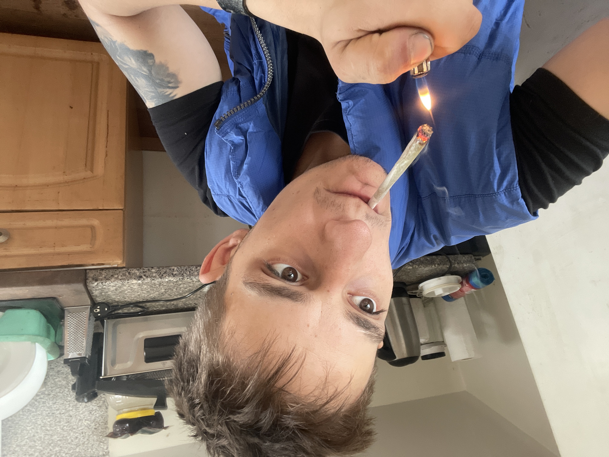 A person is shown close-up, holding a lighter to a lit cigarette. They are wearing a blue vest over a black shirt. The background appears to be a kitchen, with a countertop, cabinets, and various kitchen appliances visible. The lighting is natural, coming from an unseen window or overhead source. The person's face is turned towards the camera, and their expression is neutral.