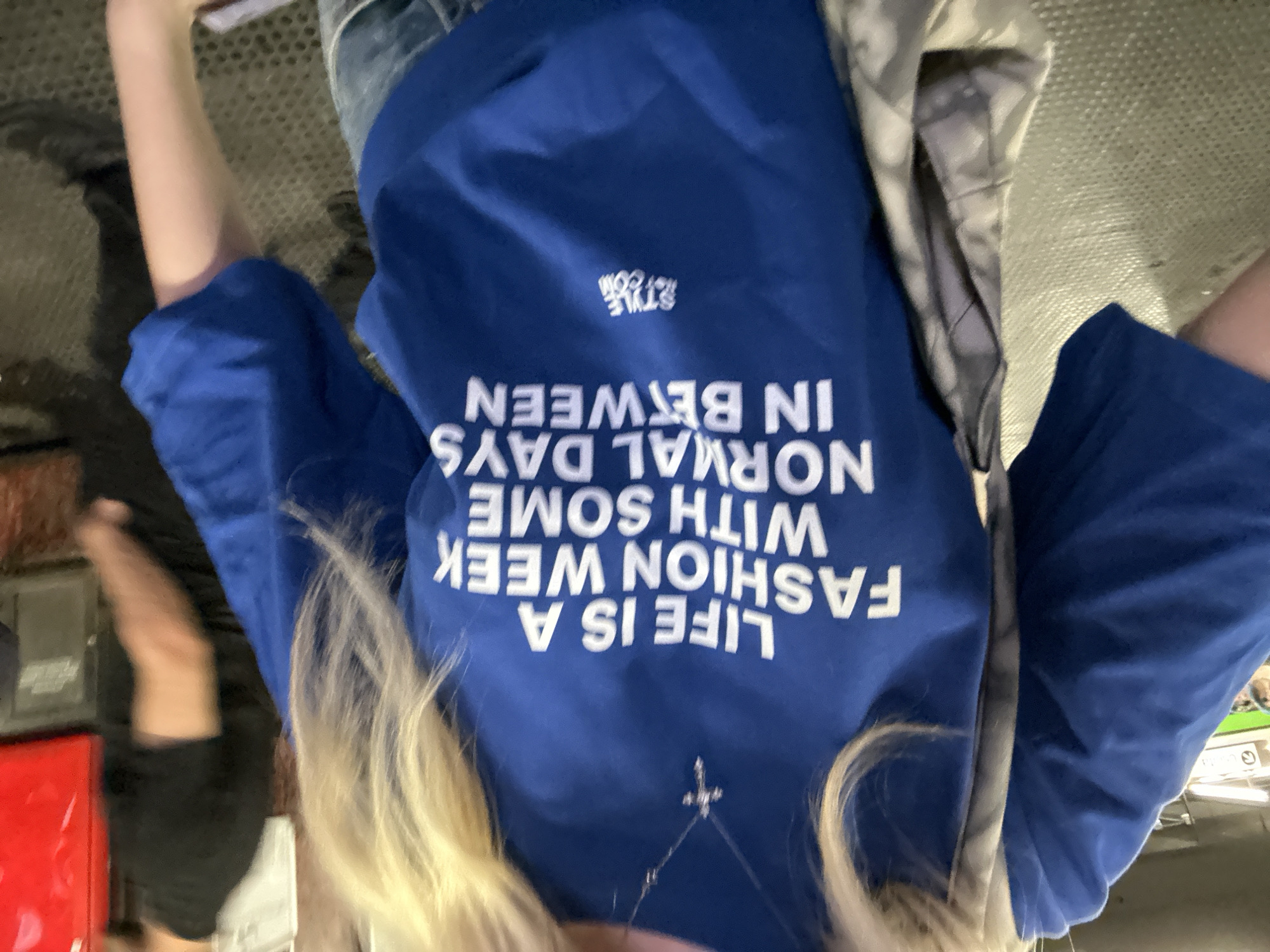 The vertically oriented image captures a person, likely female, from an overhead perspective. The subject is wearing a royal blue t-shirt with prominent white text. When correctly oriented, the text on the t-shirt reads: "LIFE IS A FASHION WEEK WITH SOME NORMAL DAYS IN BETWEEN", with "STYLE 121.COM" in smaller text above it. Long blonde hair falls over the shoulders, and a silver necklace with a small cross pendant is visible around the neck. The person appears to be wearing jeans and has a grey and white patterned jacket or bag strap draped over one shoulder.
The background shows a dark, textured floor, possibly carpet or concrete. Blurry shapes and hints of other people or objects suggest an indoor, public setting, possibly an event space. A red object, possibly a barrier or display, is partially visible in the lower-left corner. The lighting appears bright, indicative of daytime or a well-lit interior. Given the text on the shirt and the provided context, the scene is likely set during Milan Fashion Week or a similar fashion event in Milan, Italy, with the individual attending or participating.
