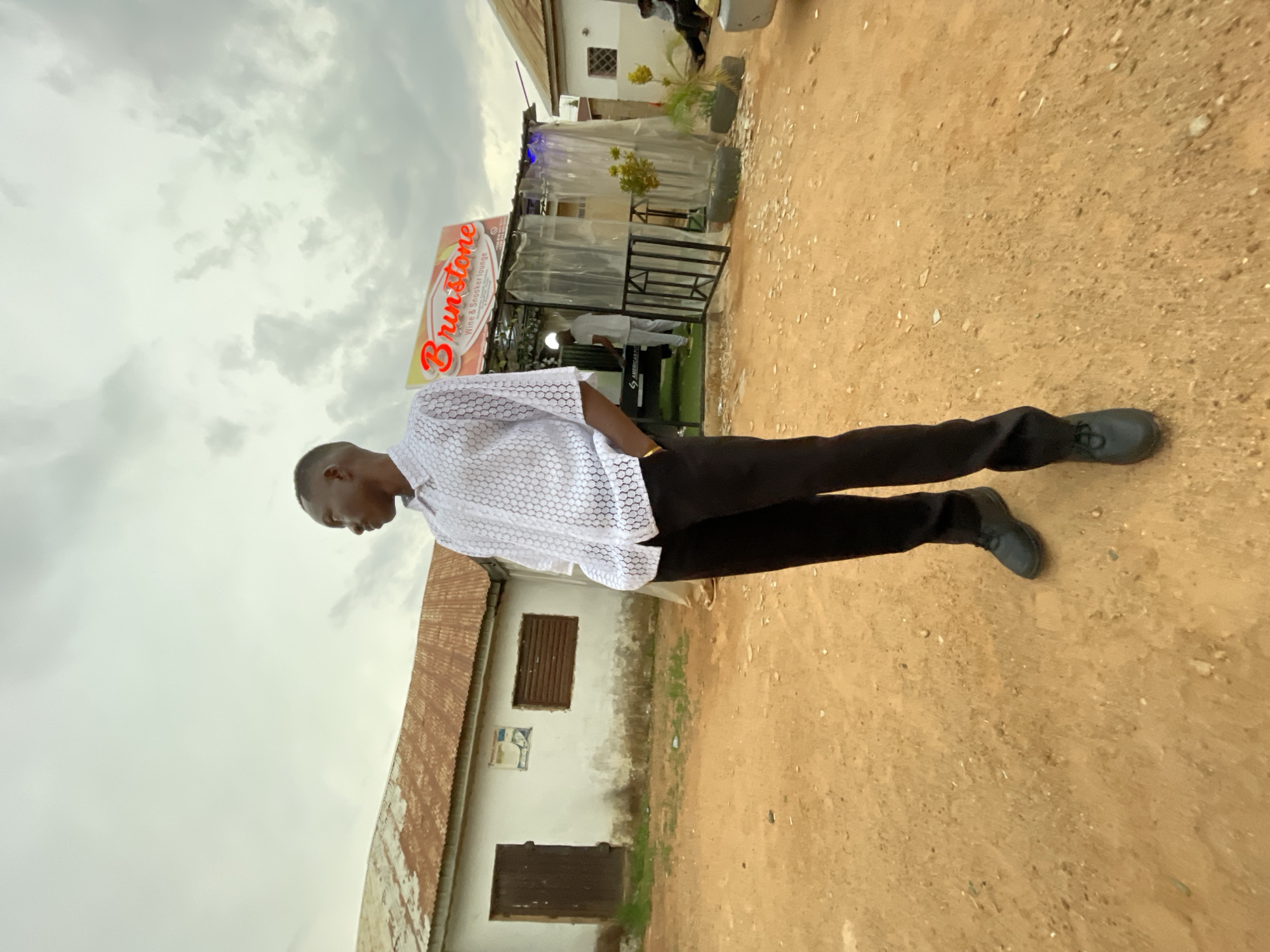 A man is standing in front of a "Brunstone" sign. He is wearing a white shirt and black pants with dark shoes. Behind him, there is a building with a brown roof. The ground appears to be dirt. The location is Jos, Nigeria, based on the sign.