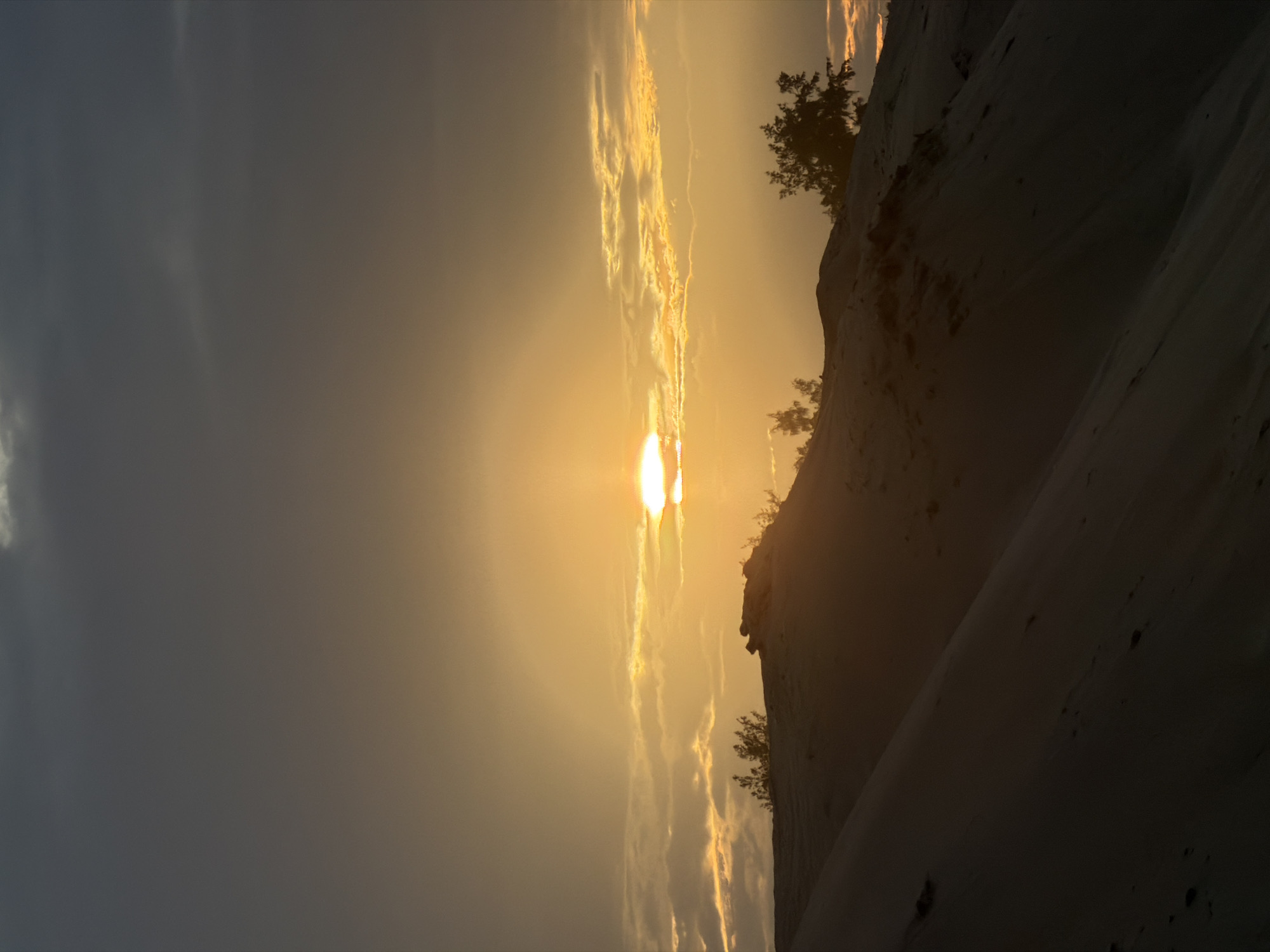 A serene sunset unfolds over sand dunes in Hoi An, Vietnam. The sun, a glowing orb, peeks through a band of clouds, casting a warm, golden light across the scene. The sky transitions from a deep gray at the top to a vibrant orange and yellow near the horizon. Silhouetted against the dramatic sky are the undulating curves of sand dunes, their surfaces textured by the wind. Sparse, dark green trees and shrubs dot the crests of the dunes, adding a touch of life to the desolate landscape. The foreground is dominated by the dark shadows of the dunes, creating a stark contrast with the illuminated sky. There are no people or animals visible in this tranquil desert-like environment.