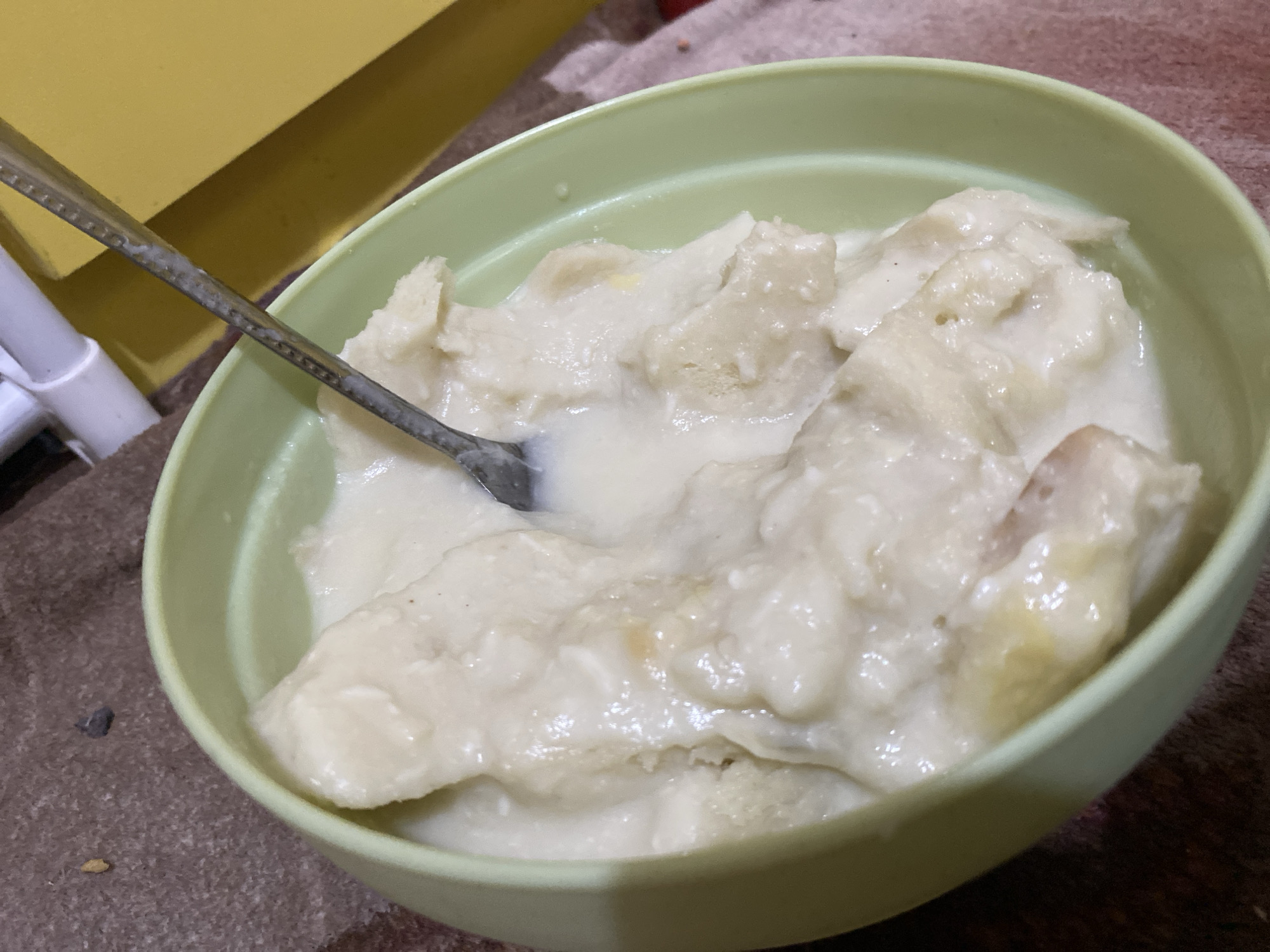 A close-up, high-angle shot shows a light green bowl filled with a creamy, white, mashed food item. A metal spoon with some food residue is resting in the bowl, partially submerged in the creamy mixture. The food appears to be composed of soft, irregular chunks within a thick, liquid base, possibly indicating a dessert or a porridge-like dish. The background is out of focus, but a yellow object is visible to the upper left, and a brown, textured surface, possibly a table or mat, occupies the lower portion of the image. The scene is set indoors, with no direct indication of the specific location within Lambaro Kueh, Indonesia, other than it is likely a domestic setting. There are no people visible in the image.