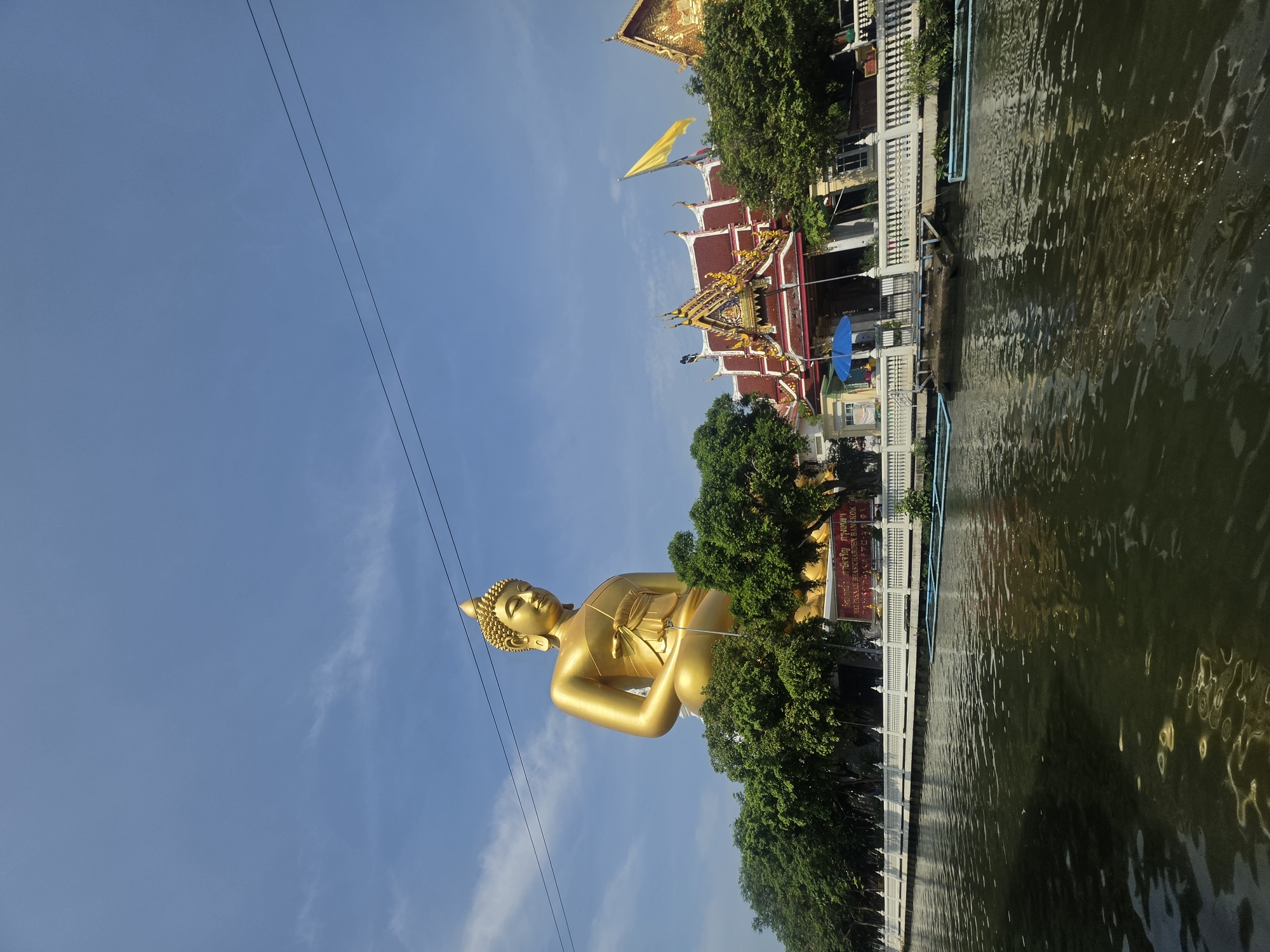 A sideways medium shot shows a golden Buddha statue sitting among trees next to a river in Bangkok, Thailand. The sky is blue with wispy clouds. Powerlines run above the statue. Behind the statue is a traditional Thai building with a red and gold roof, and a yellow flag flying above it. A white wall runs along the riverbank, with trees and buildings beyond it. The river is dark and reflects the sky and buildings.