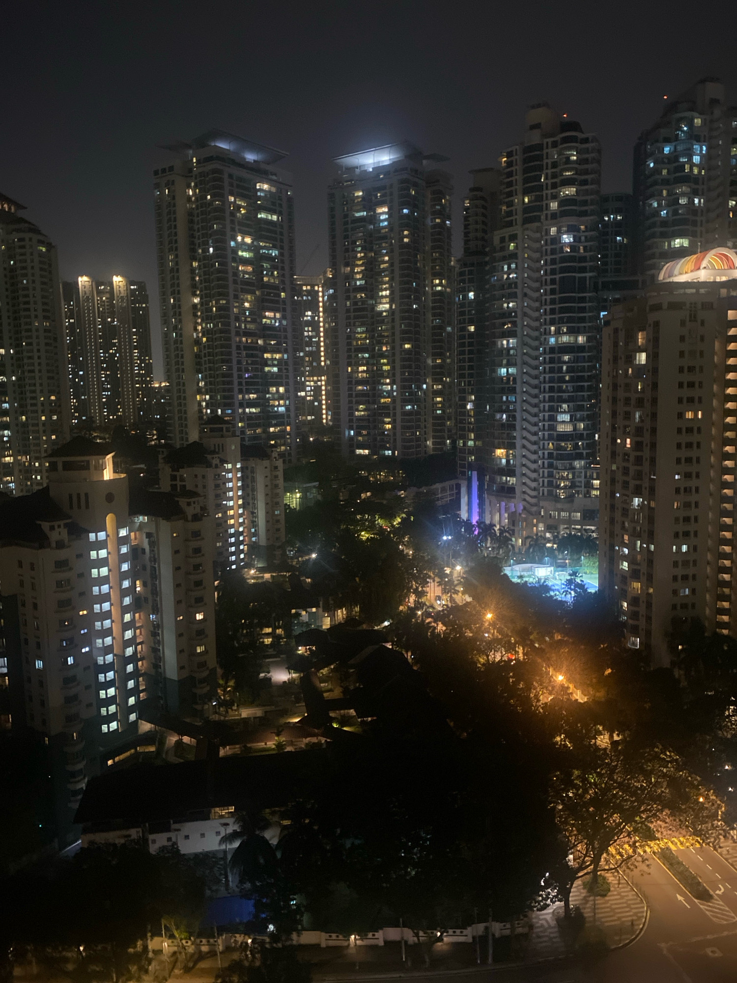 The image shows a nighttime aerial view of a densely populated urban area in Kuala Lumpur, Malaysia. Numerous high-rise residential buildings, illuminated by internal lights, dominate the skyline. The architecture varies, with some buildings featuring modern, sleek designs and others appearing more ornate. In the foreground, there are several lower-rise buildings and trees, casting shadows across the scene. Streetlights and building lights create a network of illumination throughout the cityscape. A recreational area, possibly a swimming pool and tennis court, is visible in the middle ground, lit with blue lights. The overall impression is one of a vibrant and active city at night.