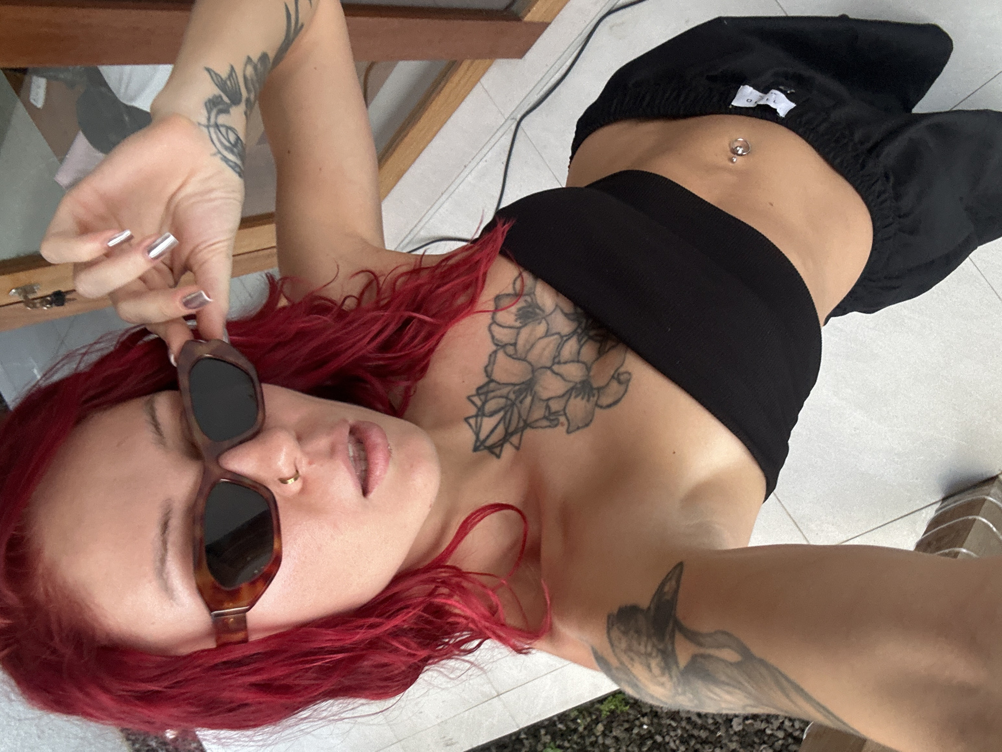 A young woman with vibrant red, long hair is depicted lying on her back, looking directly towards the camera. Her pose, with her body oriented vertically in the frame, suggests a selfie or a photo taken from above. She is wearing a black bandeau top and loose black pants, revealing her toned midriff adorned with a silver navel piercing. Brown, square-framed sunglasses are pushed up onto her forehead, and a small gold hoop nose ring is visible on her left nostril.
Her fingernails are long, rectangular, and painted a striking metallic silver. Several black ink tattoos are visible across her body: a prominent floral design, possibly peonies, on her chest/sternum, an intricate pattern on her left forearm, and a partial image of what appears to be a bird or creature on her right shoulder/upper arm.
The background consists of light-colored, possibly white or light grey, tiled flooring with visible grout lines. A black electrical cord runs across the floor. In the upper left, a wooden furniture piece is partially visible, while the lower right shows hints of outdoor greenery and bright light, suggesting an open-air or semi-outdoor setting. A small white tag with a partial brand name (possibly beginning with "OPE") is visible on the waistband of her black pants. The lighting indicates it is daytime. The scene evokes a relaxed, confident mood in a tropical environment, consistent with the location context of North Kuta, Indonesia.