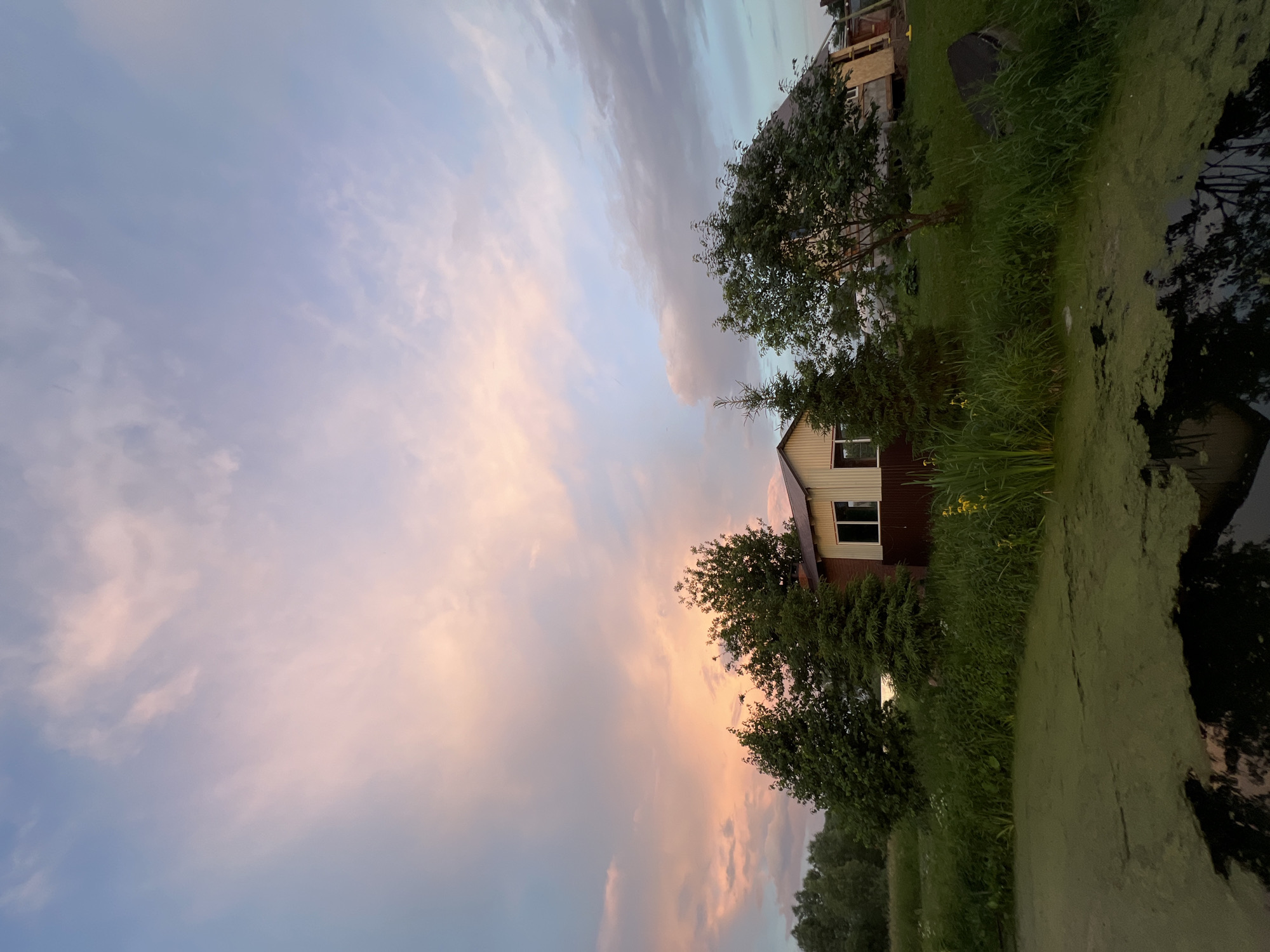 The image depicts a rural landscape at dusk in the village of Alnash, Russia. In the foreground, a body of water is covered with green algae, reflecting the sky and surrounding vegetation. A dense green embankment borders the water, with tall grasses and a few yellow flowers. Behind the embankment, a small house with a brown and white facade stands amidst trees. To the right of this house, a larger building, possibly under construction or renovation, is visible. The sky above is filled with dramatic clouds, tinged with pink and orange from the setting sun, contrasting with patches of blue. The overall scene conveys a sense of tranquil, natural beauty at the end of the day.