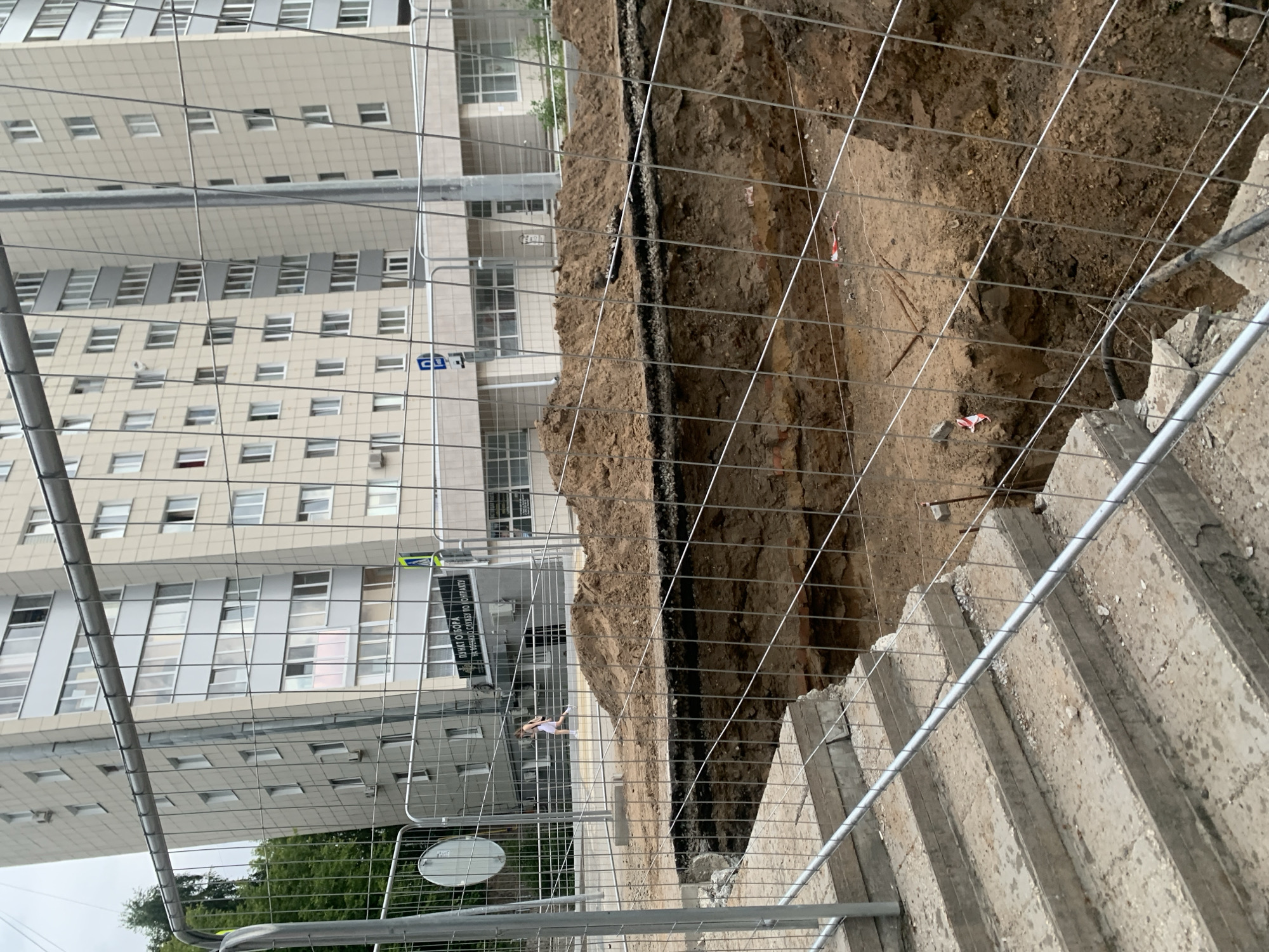 The image depicts a construction site in the city of Perm, Russia, characterized by a deep excavation in the foreground, surrounded by a temporary metal mesh fence. In the background, a modern, multi-story apartment building stands tall. The excavation reveals layers of soil and what appears to be buried brickwork, suggesting underground infrastructure work or foundation preparation. A person, possibly a construction worker or pedestrian, is visible crossing the street in the middle ground, a small pile of dirt is on top of the excavation, and a section of concrete stairs leads down towards the construction area, also partially obscured by the fence. The overall scene suggests ongoing urban development or maintenance in a residential area.