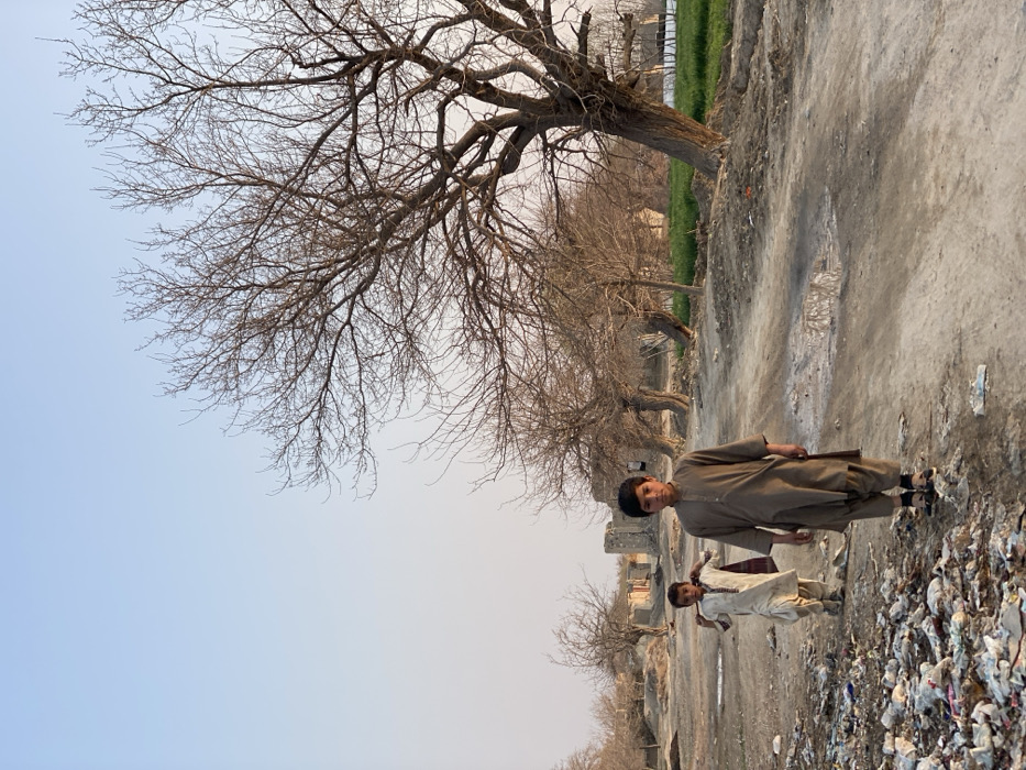The image depicts a rural or semi-urban scene in Kandahar, Afghanistan, under clear, pale sky. Two young boys are present on a dirt path.
In the foreground, a boy, approximately 8-12 years old, stands centrally, facing the camera with a neutral expression. He wears a light brown or tan shalwar kameez and dark sandals. To his immediate left, a substantial accumulation of diverse refuse, including plastic bags and other discarded materials, lines the edge of the dirt path.
Behind the first boy, slightly to the left in the mid-ground, a second, younger boy, approximately 4-7 years old, walks away from the camera. He wears a light-colored (off-white/cream) shalwar kameez and carries a dark red or maroon patterned textile over his left shoulder.
The dirt path exhibits minor undulations and features a few small puddles reflecting the sky. Along the right side of the path, a line of several deciduous trees with bare branches extends into the distance, suggesting a winter or early spring season. Beyond these trees, a patch of green vegetation is visible. In the background, to the left of the trees, dilapidated or ruined mud-brick structures stand under the pale sky. The overall light suggests daytime, possibly late afternoon.