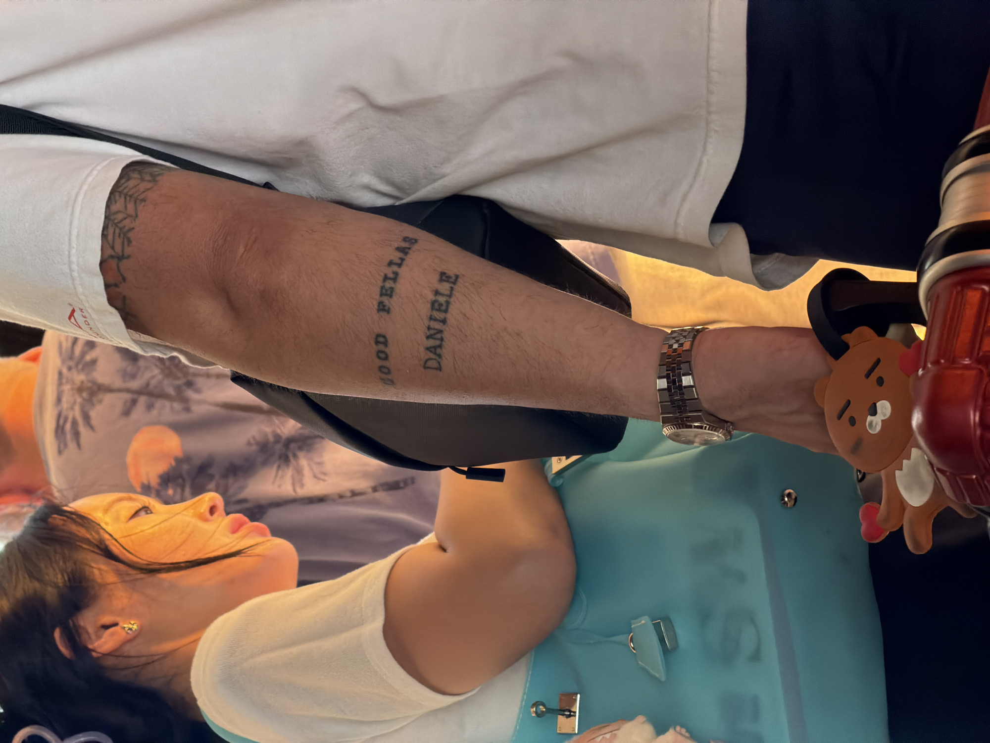 The image shows a close-up of two individuals, one of whom has tattoos on their arm. The person on the left is a woman with dark hair, wearing a white t-shirt and a turquoise bag. She has an earring in her left ear. The person on the right is partially visible, with tattoos on their forearm that read "GOOD FELLAS" and "DANIELE". They are wearing a white t-shirt and a watch on their left wrist. A black bag is slung across their shoulder. In the foreground, there are some toy-like figures. The background is blurred, with hints of patterned fabric and warm lighting, suggesting an indoor setting. The location of Baan Bang Rak, Thailand is not evident from the visual information presented in the image.