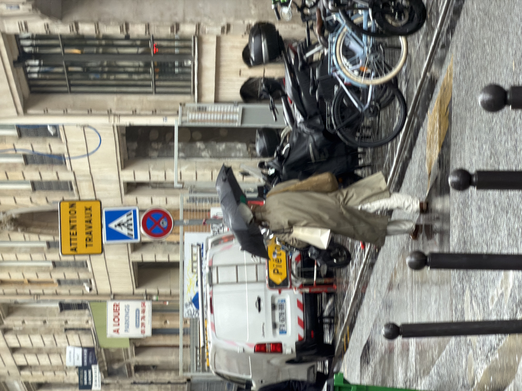 The image captures a street scene in Paris, France, on a wet day. A pedestrian, carrying an umbrella and a light-colored bag, is walking on the sidewalk beside a street with parked vehicles, including bicycles and a white van with a ladder on the back. A "Pietons" (Pedestrians) sign is visible on the back of the van. Street signs, including "Attention Travaux" (Attention Road Works) and signage for "A Louer Parkings" (Parking for Rent), are also present. The architecture of the buildings suggests an urban setting.