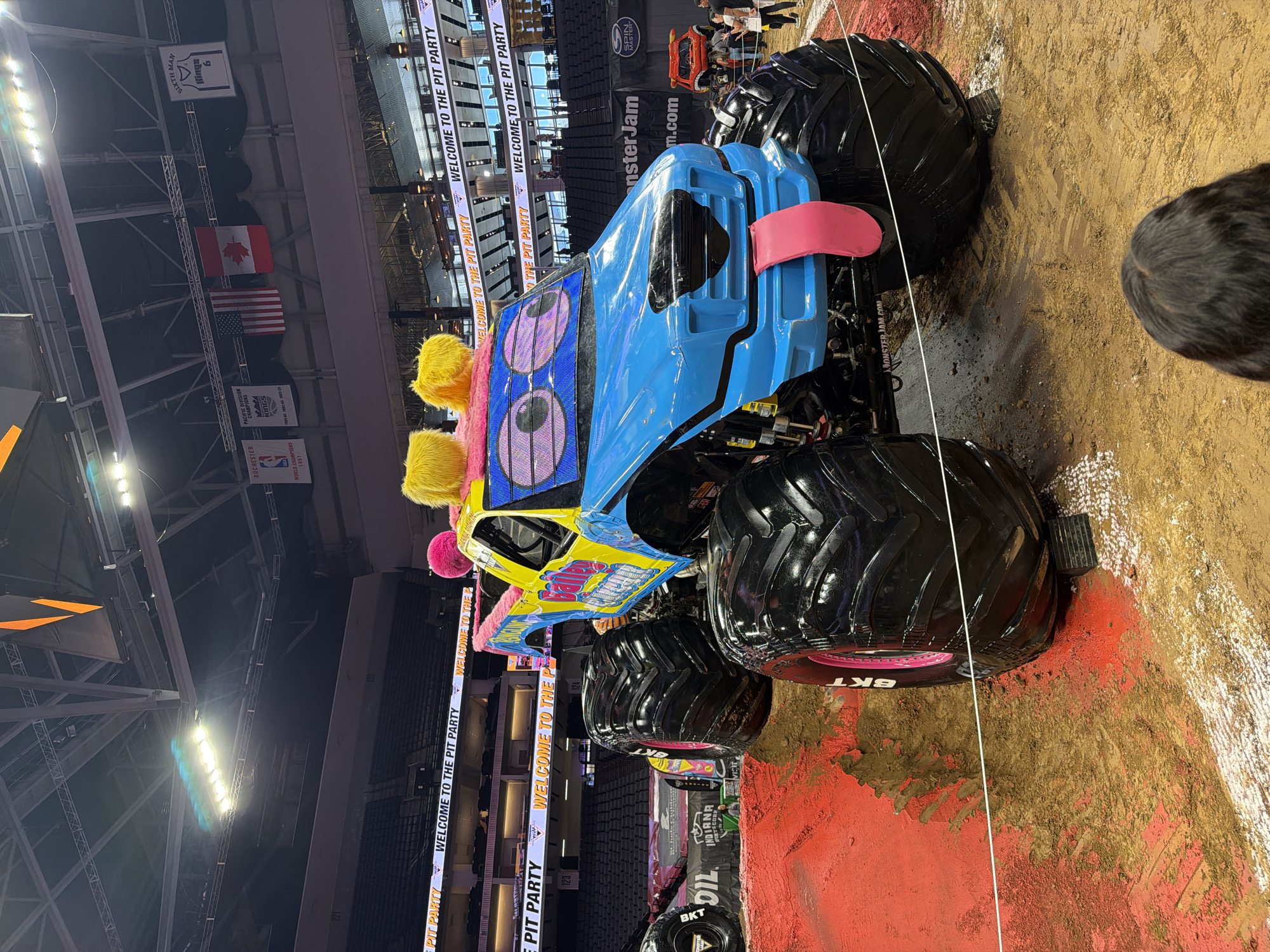 The image shows a monster truck named "Baily & Cream" on a dirt track inside an arena. The truck is blue and yellow, with large, cartoonish eyes and fluffy yellow hair on top. It has a pink tongue sticking out from its grille. The arena appears to be a sports stadium, with tiered seating visible in the background. Banners hang from the ceiling, including one that says "PACIFIC DIVISION CHAMPIONS" and flags of the United States and Canada. The words "WELCOME TO THE PIT PARTY" are displayed on screens around the arena. A red-colored track surface is visible in the foreground, with tire tracks imprinted in the dirt. Several people are visible in the background, including some standing on the arena floor and others in the stands. One person's head and shoulders are visible in the foreground, looking towards the monster truck. The overall scene depicts an event at an indoor arena.