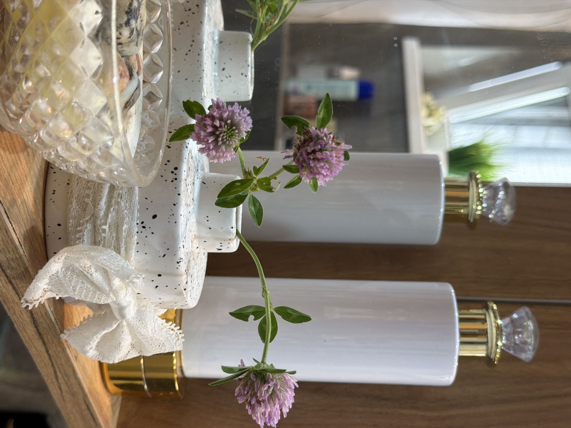 The image displays a close-up, vertically oriented view of several objects arranged on a wooden surface, likely a shelf or vanity. The primary subjects are two white cylindrical containers with gold accents and clear, faceted tops, resembling perfume bottles or decorative dispensers. Between them, a sprig of clover with pinkish-purple flowers and green leaves is artfully placed, adding a touch of nature to the scene.
To the left, a diamond-patterned glass dish holds some unclear items, possibly stones or decorative elements, partially obscured by a white lace bow tied around a speckled white ceramic object that seems to be the base of one of the cylindrical containers. The speckled pattern on the ceramic is black. A reflection of the scene is visible in a mirror or shiny surface behind the objects, showing more of the white containers and a blurred background that includes what appear to be cosmetic bottles.
The lighting suggests an indoor setting, possibly a bathroom or dressing area, with diffused light. There is no indication of the time of day or weather conditions. The arrangement feels decorative and intentional, possibly aiming for an elegant or minimalist aesthetic. No people are visible, and no text is discernable in the image. The overall mood is calm and refined.