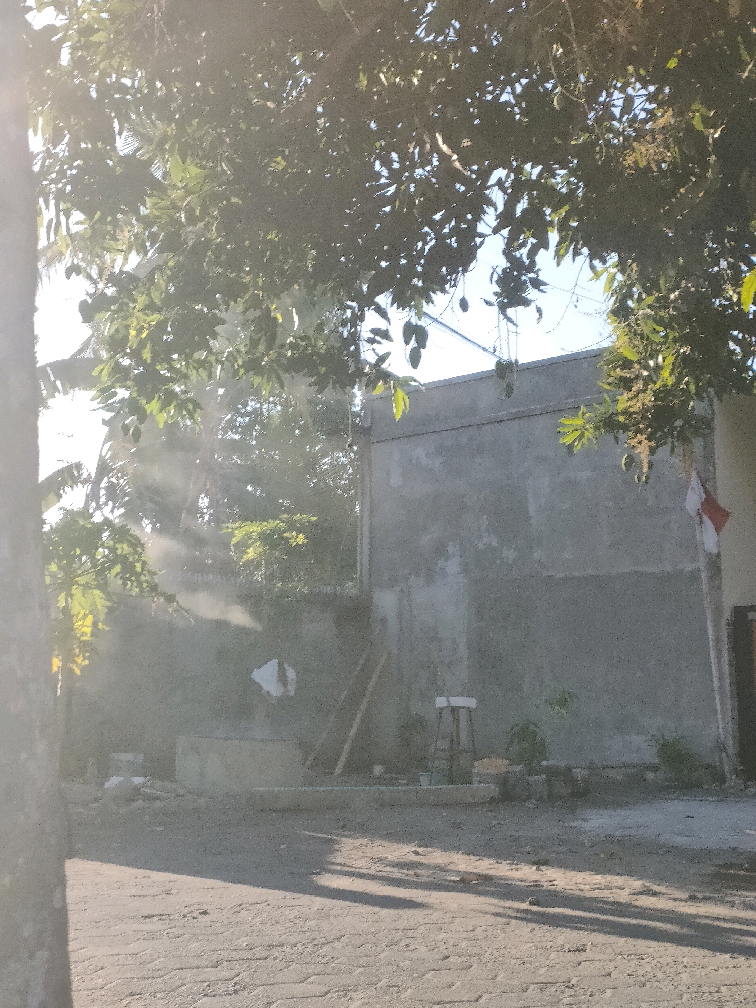The image depicts a view of a concrete wall in Kasihan, Indonesia, partially obscured by the branches and leaves of a large tree. The foreground shows a paved area with shadows cast by the tree. In the middle ground, there is a concrete structure, possibly a well or a planter, with some debris and a white bag nearby. A stool and some plants are visible against the wall. A flag, red and white, is attached to a pole near the right edge of the wall. The background shows more greenery and a clear blue sky. The scene appears to be outdoors in what could be a residential or semi-industrial area, with signs of recent construction or renovation. The lighting suggests it is daytime, possibly late afternoon due to the long shadows.