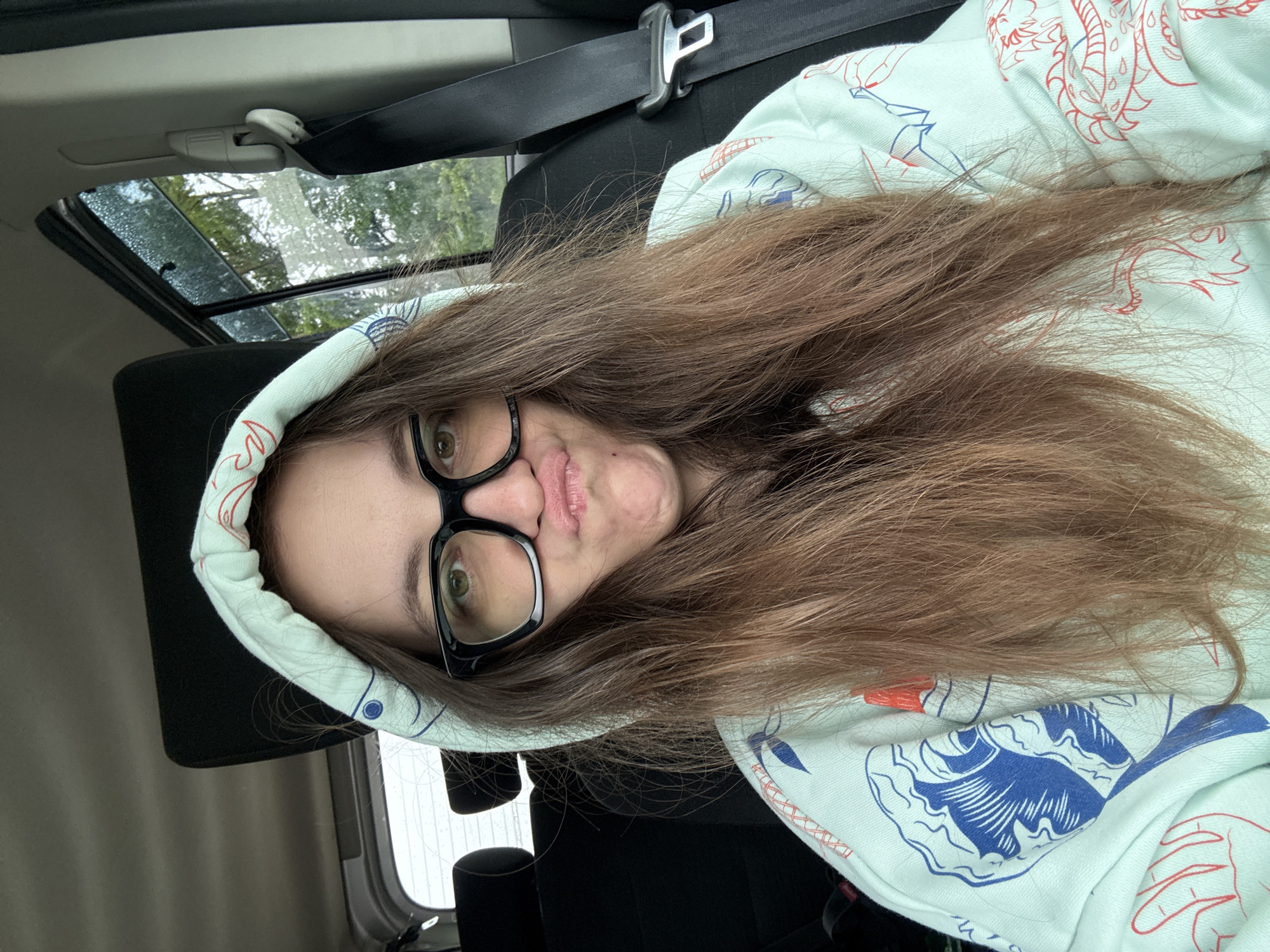 A woman is in a car, wearing a light-colored hooded sweatshirt with blue and red designs, and black-framed glasses. She has long brown hair. A seatbelt is visible across her chest. A sunroof is above her. The location Фоки, Russia is not visible in the image.