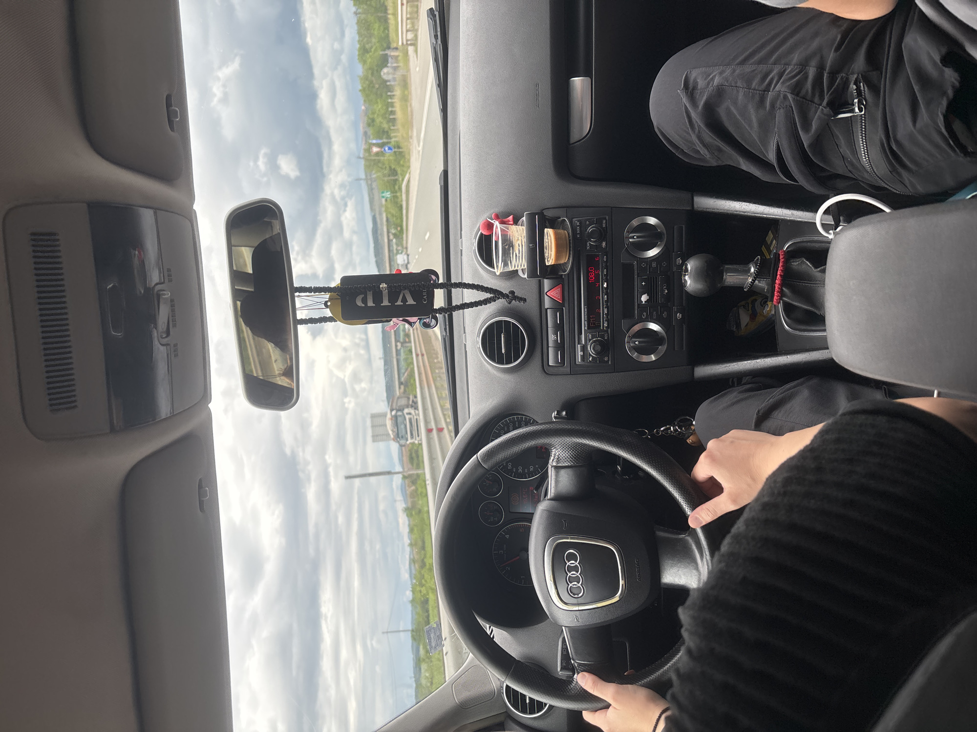 The image provides an interior view of an Audi car, rotated 90 degrees, from the driver's perspective looking out through the windshield and across the dashboard. It depicts a daytime scene with two occupants. The driver, wearing a black knitted long-sleeved top, has both hands on the steering wheel, which prominently displays the Audi logo. Next to the driver, a passenger's legs are visible, clad in black cargo pants with zippered pockets.
The car's dashboard features a central console with a radio showing "106.0 FM" and climate control knobs. A small, clear glass or cup is held by a clip attached to an air vent, with a round, cork-like object next to it. A red hazard light button is visible below the radio. A "VIP" tag, black with white lettering, hangs from the rearview mirror. The manual gear stick is visible to the driver's right, with a small snack bag or item tucked near it, and keys with a keychain rest by the passenger's side.
Through the windshield, a road or highway stretches into the distance, lined with green trees and a guardrail. A large body of water, possibly a lake or reservoir, is visible further out, beneath a cloudy yet bright sky. A truck or lorry can be seen on the road ahead. The scene is set in Hrabarsko, Bulgaria, indicating a journey through a region that includes both road infrastructure and natural landscapes.