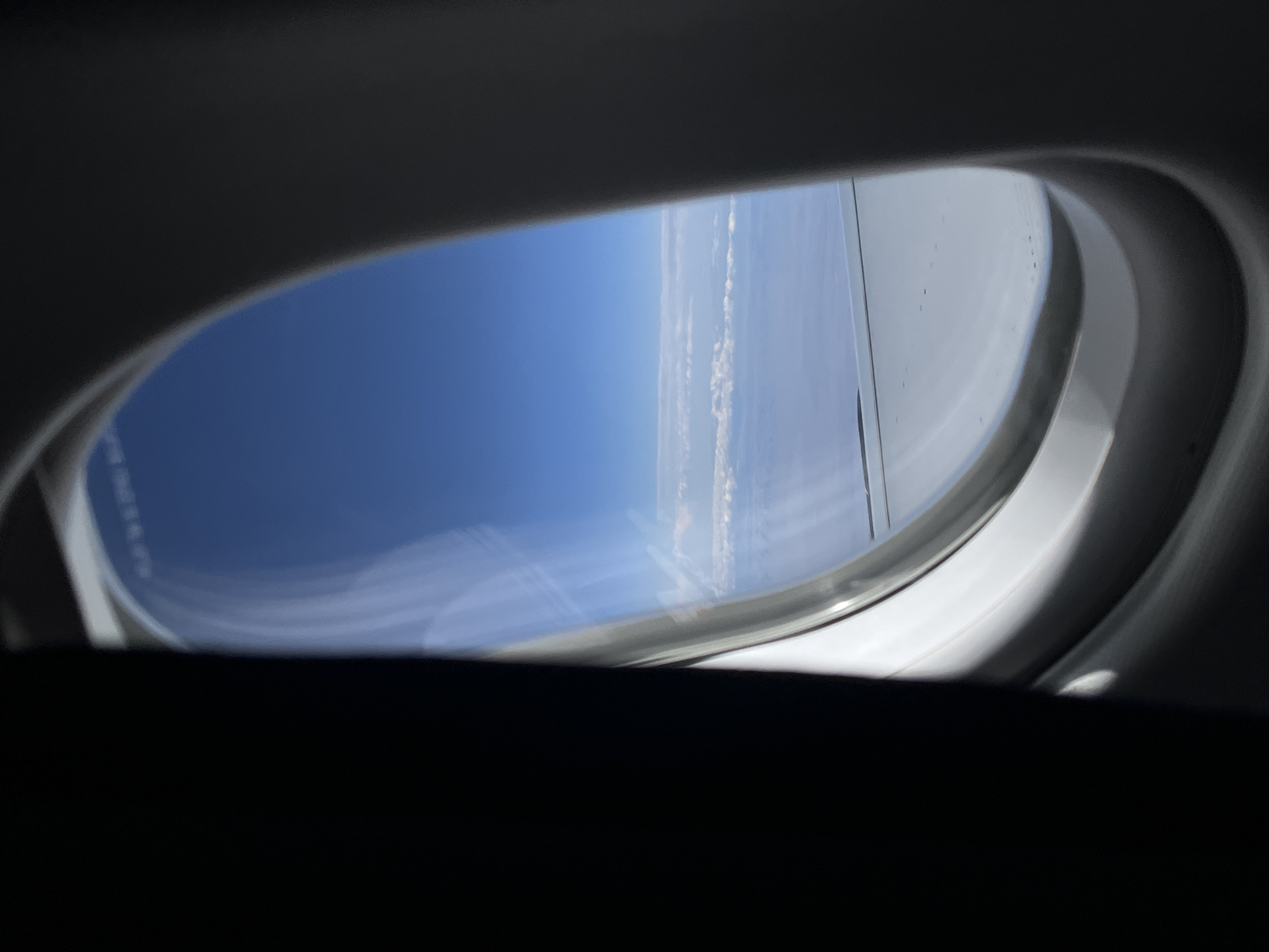 The media file captures a view from an airplane window, showcasing the vast blue sky and distant clouds from high altitude. The image is composed with the dark interior of the aircraft framing the oval-shaped window, which acts as the main viewport.
Through the window, a bright, clear blue sky dominates the upper and left portions of the visible exterior. Along the right side, an airplane wing is partially visible, indicating the aircraft's movement. Below the wing and stretching across the horizon, a layer of thin, wispy white clouds can be seen. The clouds appear somewhat scattered or in lines, suggesting a relatively clear sky above them.
The bright sunlight illuminating the sky and clouds indicates that the image was taken during daytime. There are no people visible in the frame, and the focus is entirely on the aerial landscape. Faint, blurry text, possibly a label or instruction, is barely discernible on the window's surface, but it is illegible. The setting implies active flight over a geographic area, with the provided context being Tsagaanchuluut, Mongolia. The scene evokes a sense of travel and the expansive beauty of the sky from above.