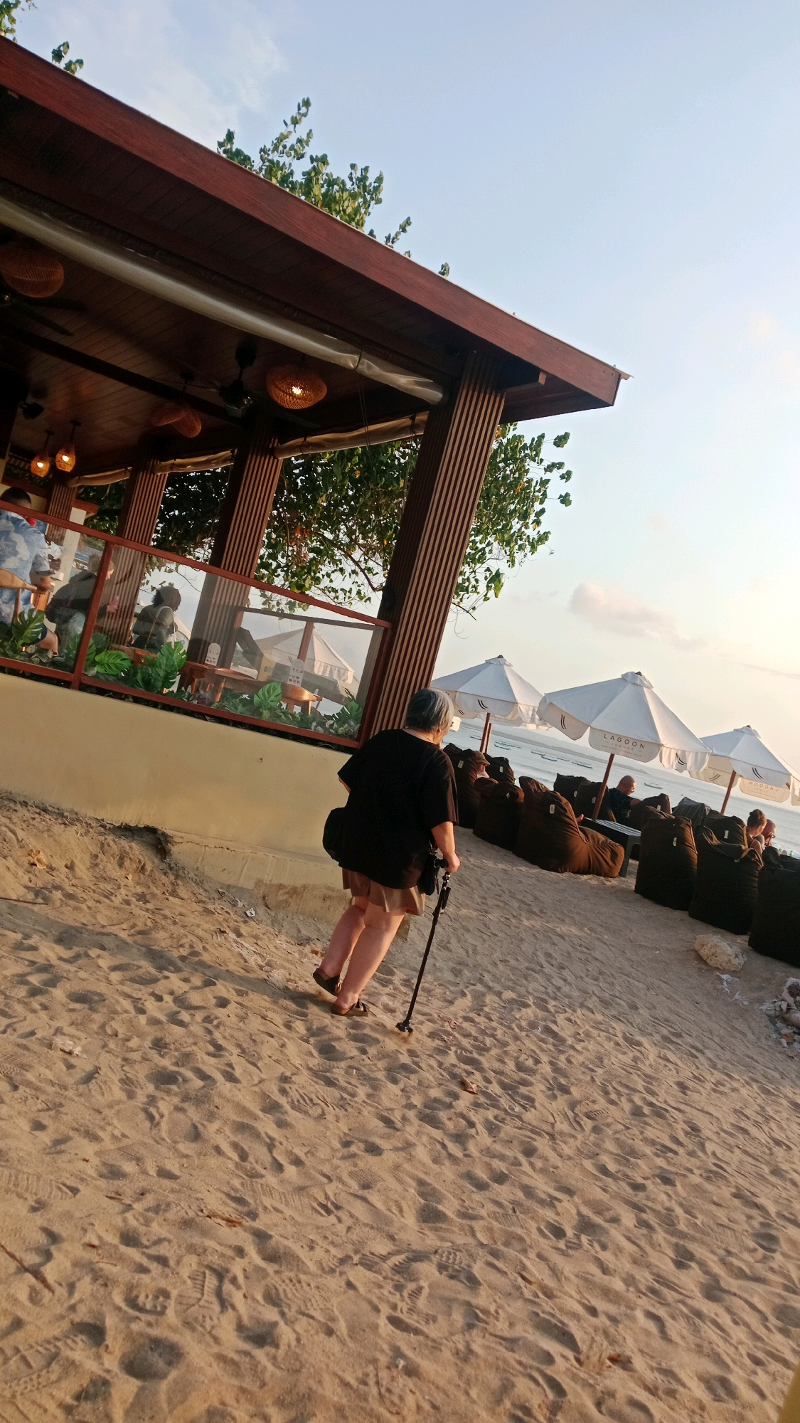 The image captures a person walking on a sandy beach in Kuta, Indonesia, near a beachfront establishment. The person, seen from the back, is an adult with graying hair, wearing a dark t-shirt, a light-colored skirt, and sandals, and is using a walking stick. They are walking away from the viewer, towards the ocean and a row of white umbrellas and large beanbag chairs. The establishment has a wooden roof and vertical wooden beams, with glass partitions showcasing plants and seating areas inside. The sky is clear and bright, indicating daytime. The sand is marked with footprints, and the overall lighting suggests it is either late afternoon or early morning.