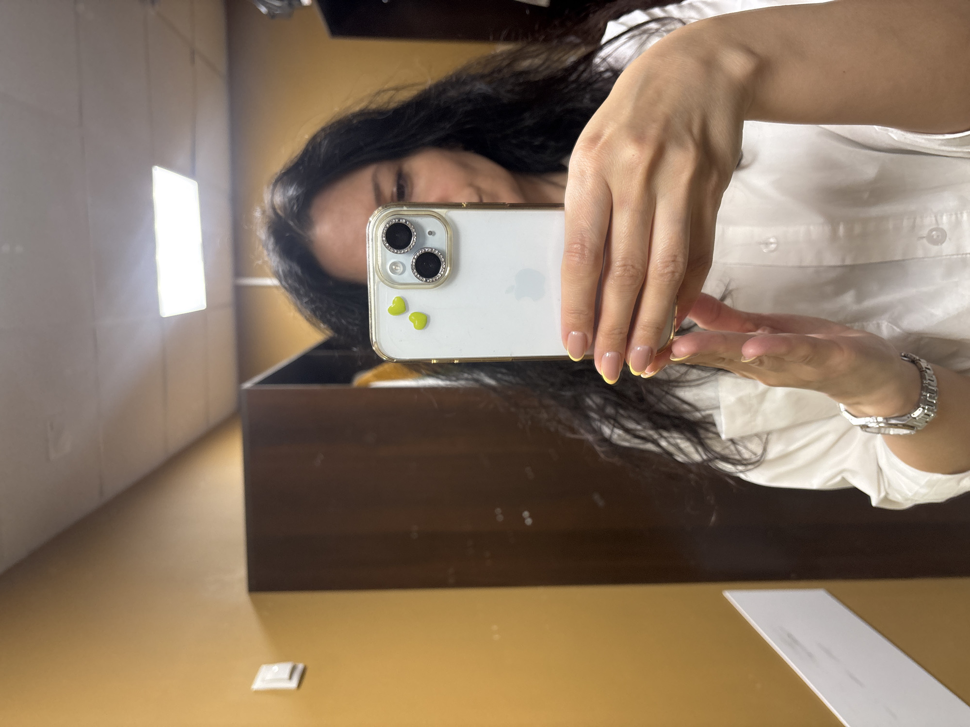 A mirror selfie shows a woman holding up a white smartphone with a clear case decorated with two green heart stickers near the camera lenses. Her face is partially visible behind the phone. She is wearing a white button-down shirt and a silver wristwatch on her left wrist. The reflection reveals a room with dark wood furniture, a light-colored floor, and a bright rectangular light source in the upper left corner. A white rectangular object lies on the floor in the lower right. There is no information to suggest the location is in Chaikovsky, Russia.