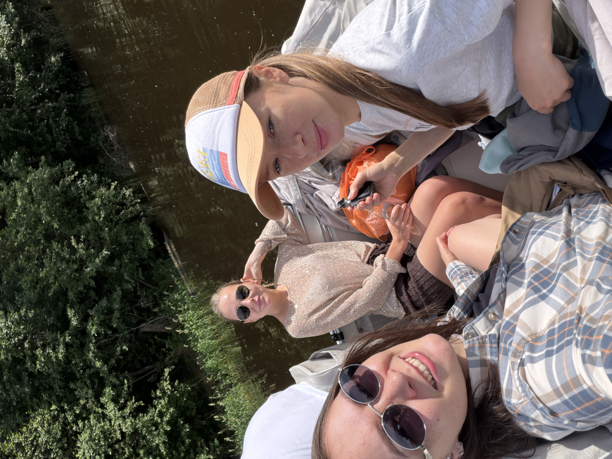 Three young women are seated in a boat on a body of water, likely a river or lake, surrounded by lush green trees and vegetation. The woman in the foreground is taking a selfie, smiling and wearing sunglasses and a plaid shirt. To her right, another woman is looking towards the camera, wearing a white and brown trucker hat and a grey t-shirt, holding a black object in her hand. Behind them, a third woman with blonde hair and sunglasses is seated, wearing a sparkling beige top and a brown skirt. An orange bag is visible between the two women in the background. The water is dark and reflects the trees. The scene appears to be a recreational outing in a natural setting. The location is Chaikovsky, Russia.
