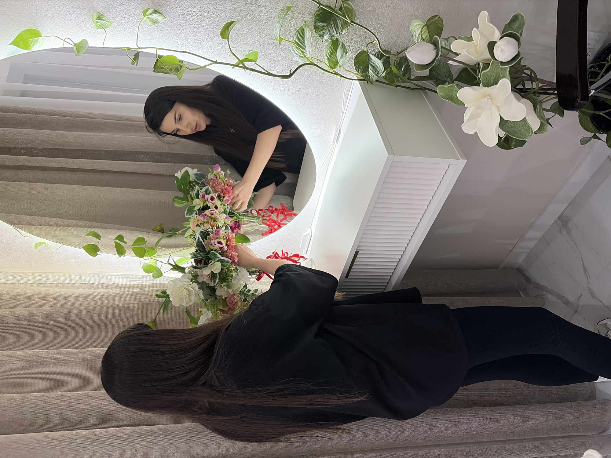 The image depicts an indoor scene in Budva, Montenegro. An adult female with long, dark hair, wearing a black oversized t-shirt and black leggings, stands with her back partially to the viewer, facing a large, oval-shaped mirror with integrated backlighting. Her reflection in the mirror shows her hands actively arranging a bouquet of artificial flowers within a clear glass vase. The vase is adorned with several red ribbons tied around its neck and contains a mix of white, pink, and green artificial blossoms and foliage.
Beneath the mirror is a white, wall-mounted console table featuring a ribbed or fluted front drawer panel. A green leafy vine trails downwards along the right side of the mirror and the adjacent wall. In the lower right foreground, a dark pot holds an artificial plant displaying large white flowers and buds. The background includes light brown, textured curtains drawn across what appears to be a window on the left. The walls are light-colored, and the floor is covered with white marble-patterned tiles.