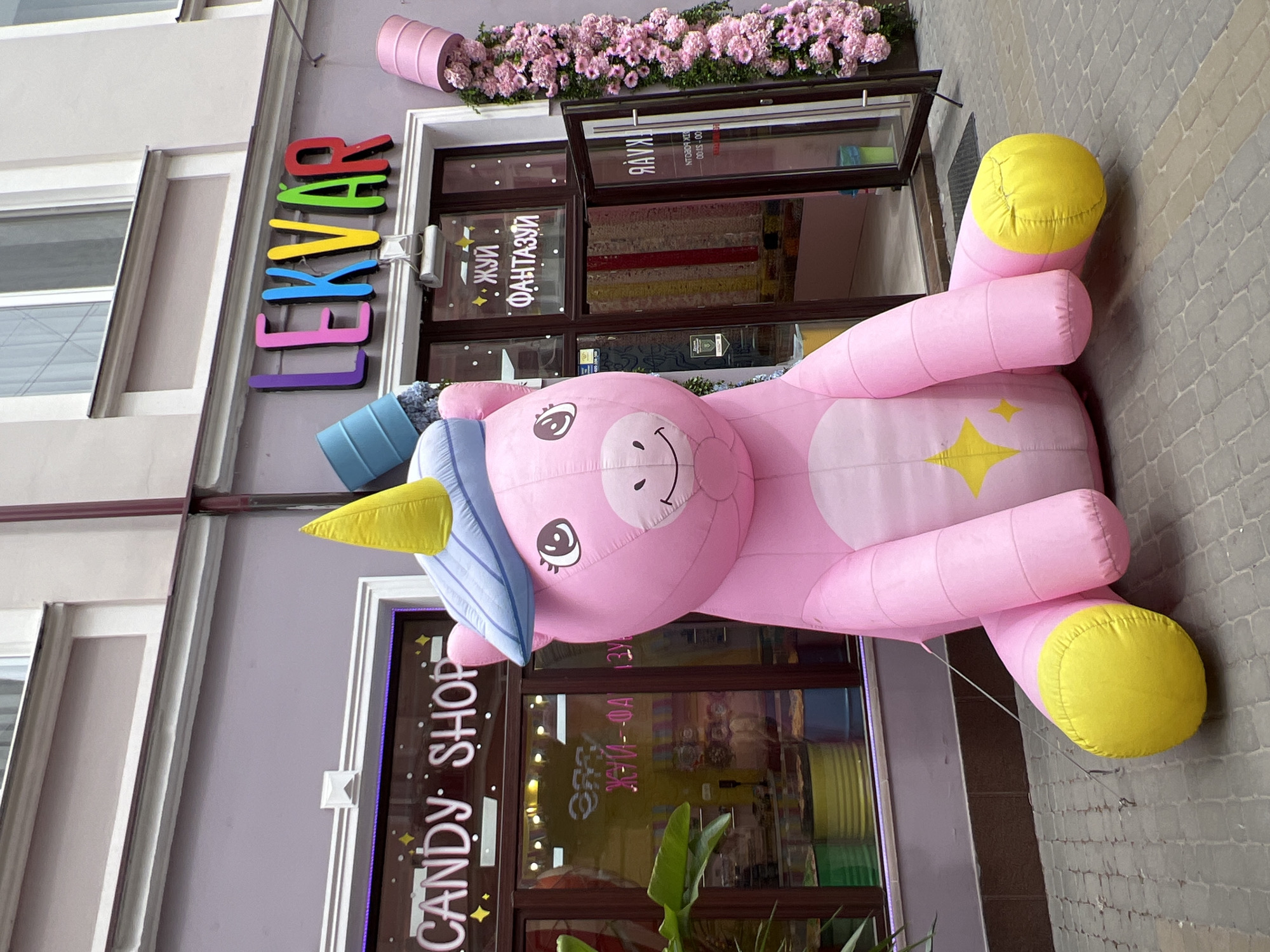 Here is a forensic description of the image:
The image shows a storefront with the name "LEKVAR" in colorful, individual letters above the doorway. Underneath is a smaller sign with the words "ЖУЙ ФАНТАЗУЙ" (translated "Chew Fantasy"), written in Ukrainian. To the left of the doorway, vertically, reads "CANDY SHOP". A large, inflatable pink unicorn lies on the sidewalk outside the shop. The unicorn has a yellow horn and feet, and a blue hat. An awning above the doorway is decorated with pink flowers. Reflections in the shop windows show the interior contains colorful displays. The location is in Ternopil, Ukraine.