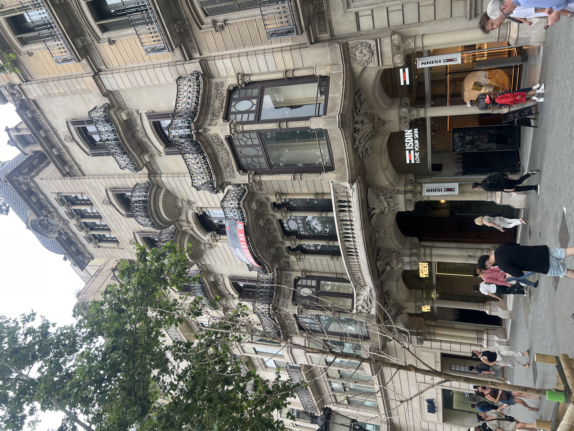 This image captures a street view of Passeig de Gràcia in Barcelona, Spain, a well-known avenue lined with modernist architecture. In the foreground, a mature tree with dense green foliage partially obscures the upper floors of an ornate building. The building itself is constructed of light-colored stone and features a multi-story facade with numerous balconies supported by decorative ironwork. Several windows are visible, some with wooden shutters.
On the ground floor of the building, a retail establishment with signage for "ISDIN LOVE YOUR SKIN" is prominent, featuring large glass storefronts and a modern interior. Two flags with the ISDIN logo hang outside the entrance. A few people are seen walking on the sidewalk in front of the store, including a man in a red and white striped shirt and blue jeans, a woman in a red dress and a patterned headscarf, and a woman wearing glasses. To the left of the ISDIN store, a store with "ONITSUKA TIGER" signage is visible.
Further down the street, other pedestrians are walking, and some shop entrances are apparent. The street is paved with cobblestones. The overall impression is of a bustling urban environment with significant architectural heritage.