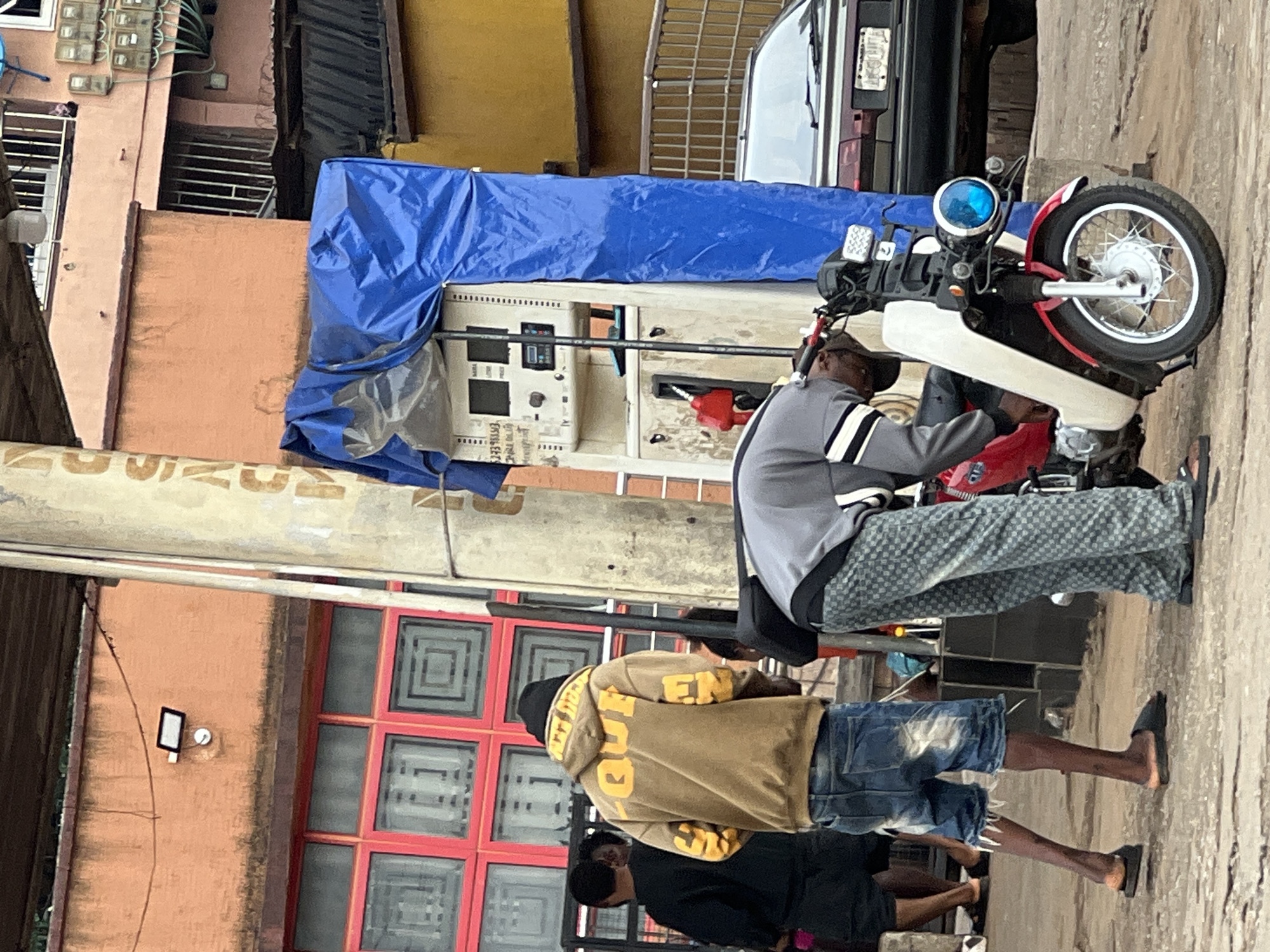 The image depicts a street scene in Nkpor, Nigeria, near a gas station. In the foreground, a red motorcycle is partially visible, with a person in a gray jacket and patterned pants attending to it. To the left, a person in a brown hooded sweatshirt and ripped denim shorts is standing. Behind them, other individuals are present, some with their backs to the camera. A gas pump, covered with a blue tarp, is a prominent feature of the scene. A dark-colored car is parked in the background. The environment suggests an urban setting with buildings and a paved ground.