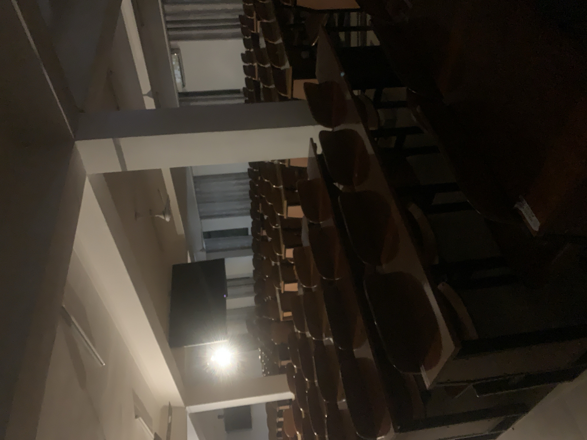 The image shows an empty lecture hall with rows of chairs and tables facing a blank screen. The hall is lit by ceiling lights, and the walls are a light color. The location is in Dar es-Salaam, Tanzania.