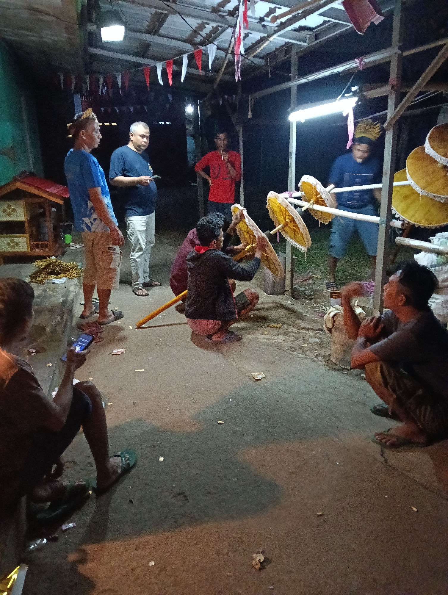 The image depicts an outdoor gathering at night in Tasikmalaya, Indonesia, likely a community event or preparation for one. Several men are present in a dimly lit area with a simple wooden structure overhead, adorned with red and white flags. Some men are standing, observing, while others are engaged in activities related to what appear to be traditional umbrellas or decorative items. Two men are crouched near a collection of these yellow, circular objects, which are mounted on wooden poles. One man, wearing a dark shirt and shorts, is actively holding and adjusting one of these objects. Another man, also crouched, appears to be looking at something in his hands, possibly related to the preparation. A few more men stand in the background, one in a blue shirt and patterned shorts, another in a black t-shirt, and a third in a red shirt. A small wooden structure with a red roof is visible to the left, and loose papers and what may be cigarette butts are scattered on the concrete ground. The overall atmosphere suggests a casual, communal activity taking place outdoors.